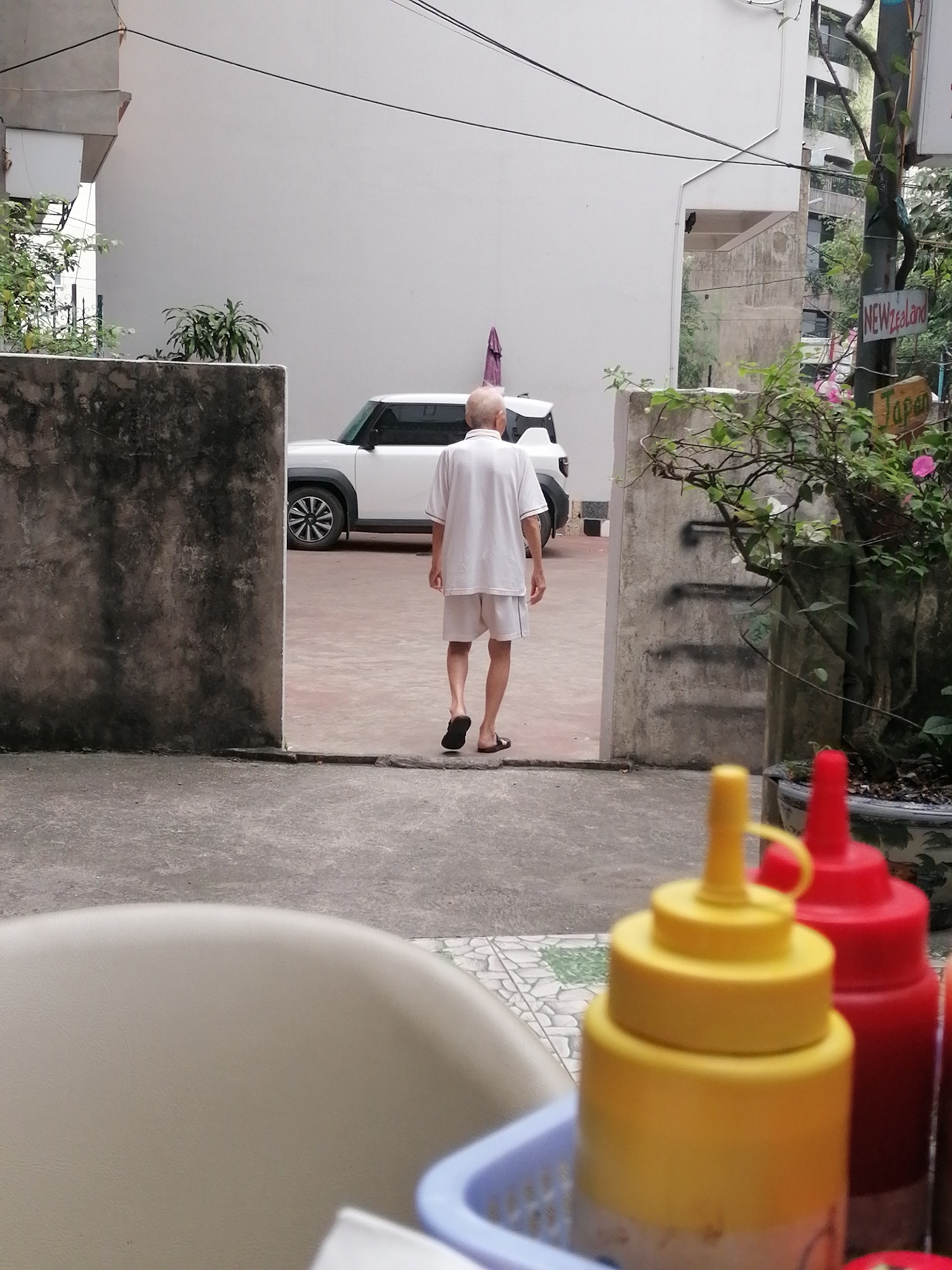 In Bai Chay Ward, Vietnam, the image shows a courtyard flanked by concrete walls on either side, creating an entrance. A white SUV is visible in the courtyard. An elderly man, wearing a white shirt and shorts, walks away from the camera towards the vehicle. To the right of the entrance, a plant in a large pot is positioned near a sign indicating "New Zealand" and "Japan." In the foreground, partially obscuring the view, are condiments: yellow mustard and red ketchup bottles. A light-colored chair back is also partially visible.