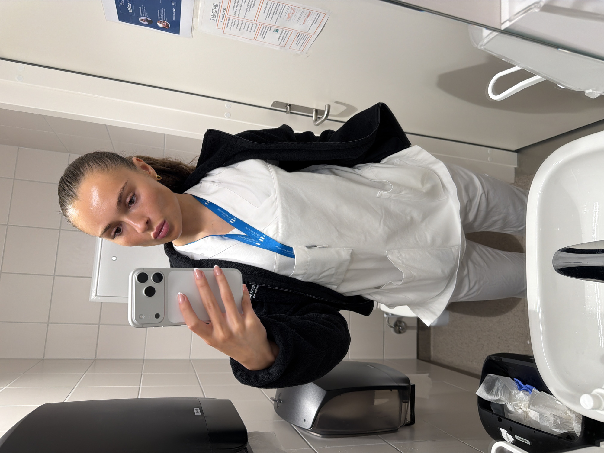 A young woman with brown hair pulled back in a ponytail is taking a selfie in a bathroom. She is wearing a white medical scrub top and white pants, and a black zip-up fleece jacket. She has a blue lanyard around her neck with a white card attached. She is holding a silver smartphone in her right hand, angled to capture her reflection. The bathroom has white tiled walls and a gray speckled floor. To her left is a black paper towel dispenser and a black waste bin. To her right is a white door with a silver handle and a poster on it. The sink area is visible in the foreground. The image is taken from a slightly low angle.