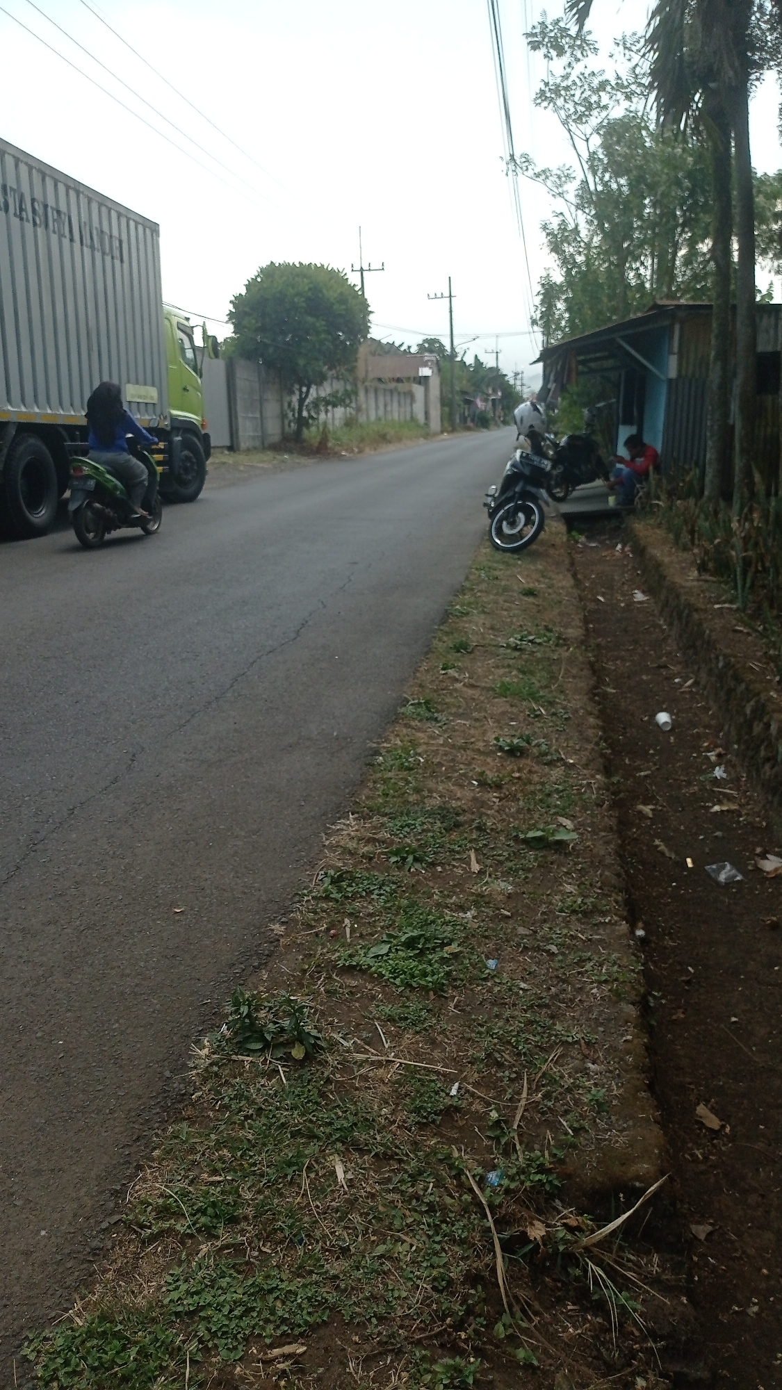 In Bedali, Indonesia, a person on a motorcycle travels on a paved road next to a large grey shipping container truck. On the right side of the road, there is a drainage ditch filled with debris. Further down the road, two motorcycles are parked. A small blue and grey structure with a corrugated metal roof is situated beside the road, with two individuals near the motorcycles. Trees line the background, and utility poles and wires are visible against the overcast sky.