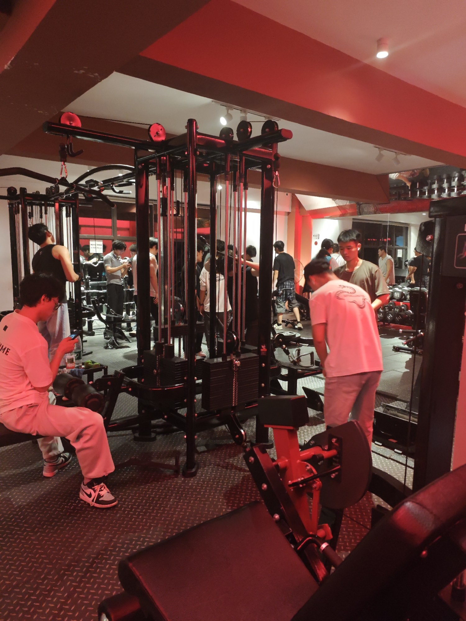 The image captures an indoor gym scene in the Chongqing High-tech Zone, China, characterized by a dominant red ambient lighting. Approximately eight to ten men are visible, engaged in various activities.
In the foreground to the left, a man in a light-colored t-shirt and sweatpants sits on a bench, looking at his smartphone. His white and black sneakers are visible. On the bench next to him, a green water bottle is placed. To the right, another man in a light t-shirt and grey pants stands next to gym equipment, facing away from the camera. A large, black multi-station cable machine with weight stacks occupies the center of the frame, around which several men are gathered. One man in a black tank top stands with his back to the camera near the cable machine, while another in a light grey polo shirt appears to be holding a device, possibly a phone or camera, and looking at it. Other individuals are seen further back, some reflected in large mirrors that line the walls, interacting or waiting near dumbbell racks and other exercise machines.
The red lighting creates an intense and somewhat dim atmosphere, with some white ceiling spotlights noticeable but overshadowed by the red hue. The floor is covered with a textured black rubber matting, common in gyms. Red elements are integrated into some of the exercise equipment, such as parts of the leg press machine in the foreground right and details on the cable machine.
Visible text includes "ME" on the back of the white t-shirt worn by the seated man in the foreground. A stylized white design or logo is on the back of the t-shirt of the man in the right foreground, turned away from the camera. Some blurred Asian script, likely Chinese characters, is reflected in a mirror in the middle-left background, possibly part of a gym notice or rules. The overall activity suggests a social gym environment where people are working out, observing, resting, or conversing. The artificial lighting implies it could be evening or night.