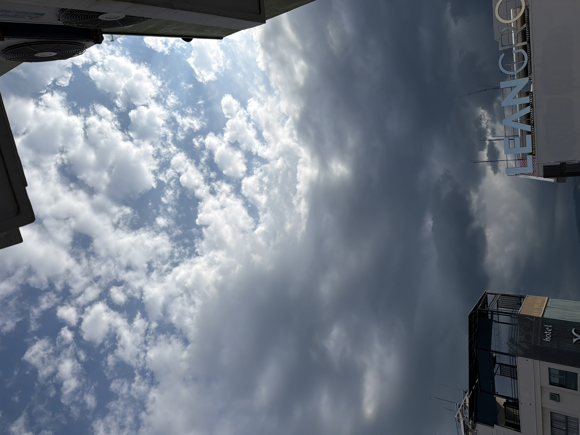 The image depicts a view from a high vantage point, looking upwards towards a sky with a dramatic contrast between bright, fluffy white clouds and darker, heavier storm clouds. The sky takes up the majority of the frame, with a small portion of buildings and air conditioning units visible at the edges. In the lower left quadrant, a portion of a modern building with glass railings and a rooftop terrace is visible. The word "hotel" is visible on the side of this building. In the lower right quadrant, another building with large, illuminated blue letters spelling out "LEAN GRC" is present. The overall impression is of a dynamic sky with a looming weather change over an urban setting. The context of Nha Trang Ward, Vietnam, suggests a coastal city, which may influence the types of clouds and weather patterns observed. However, there are no specific visual cues in the image to confirm this location beyond what is provided.