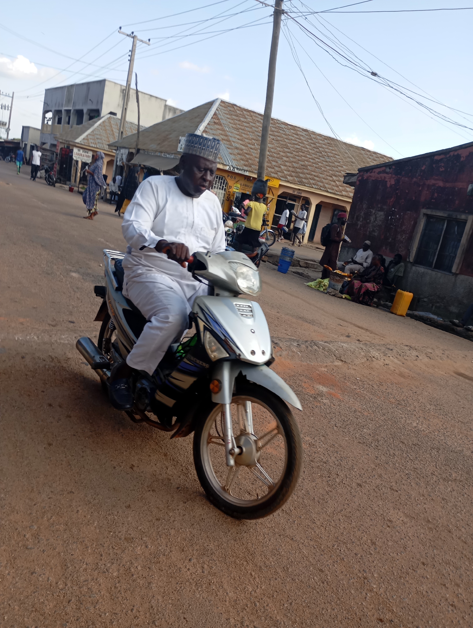 A man in white traditional attire and a cap rides a silver motorcycle on a dusty road in Shendam, Nigeria. He is in the foreground, facing slightly away from the viewer. Behind him, on the left side of the road, are several buildings with corrugated iron roofs. There are people walking and standing in front of these buildings. On the right side of the road, some people are sitting down, seemingly interacting near a stall with produce. A yellow jerrycan is visible on the right. Power lines and a utility pole are overhead. The ground is a reddish-brown dirt road.