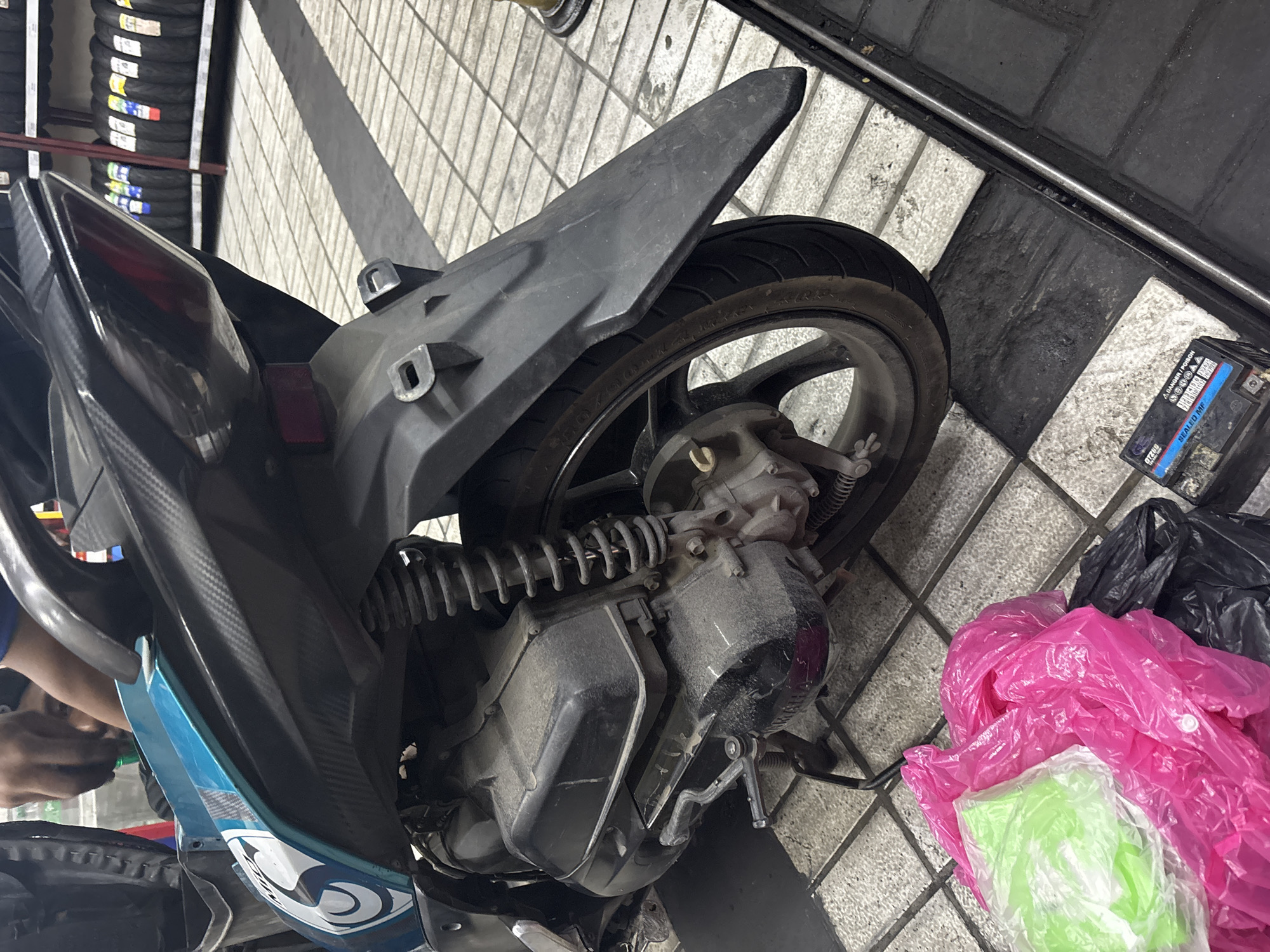 In North Jakarta, Indonesia, a close-up, eye-level shot captures the rear section of a teal and black scooter, with a person's hands working on the engine area. The scooter is positioned on a checkered tiled floor, with a shadow cast across it. To the left, several plastic bags, one bright pink and another translucent green, are piled on the floor. In the foreground, a small black battery with a warning label sits on the tiles. In the background, shelves lined with tires are visible, suggesting a workshop or store setting.