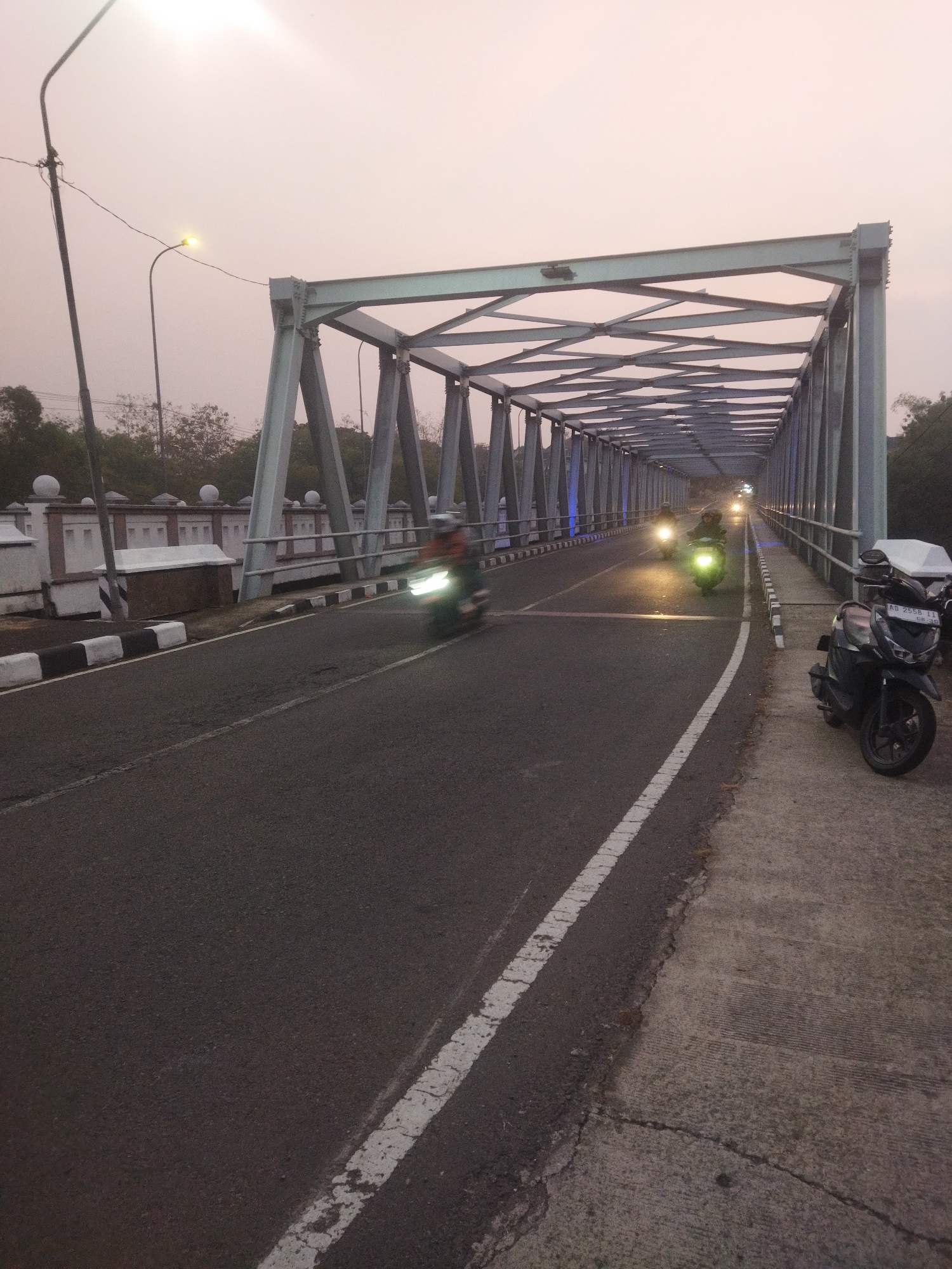 The image depicts a road bridge in Wonogiri, Indonesia, during what appears to be twilight. A metal truss bridge spans across the scene, with its structure dominating the upper half. The road surface is asphalt, marked with white lines, and it curves slightly towards the right. On the right side of the road, a concrete sidewalk is visible, and a parked black motorcycle is positioned there. Several motorcycles are in motion on the bridge, their headlights illuminated, creating streaks of light due to motion blur. Some of these motorcycles have riders, identifiable by their helmets. In the background, trees and some structures are visible. The sky is overcast with a pale pinkish hue.
