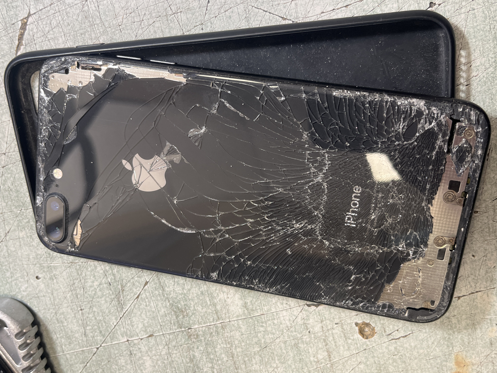 The image shows a heavily damaged black iPhone with a cracked back. The cracks are extensive and cover most of the surface. A black phone case lies partially underneath the iPhone. The background is a textured, light-colored surface. The iPhone is labeled "iPhone" and has an Apple logo in the center. The scene is indoors, but the specific location is unknown. No people are visible.