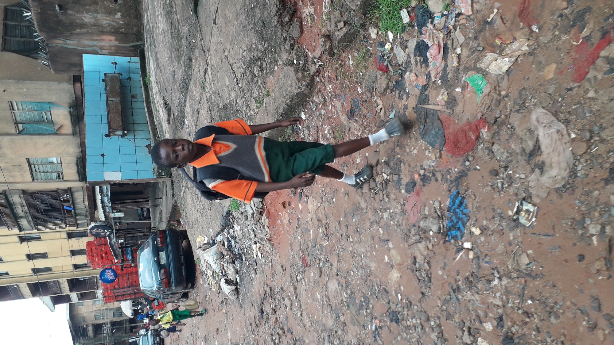 The image depicts a young person, likely a student, standing on a dirt path littered with trash in Onitsha, Nigeria. The individual is wearing a collared shirt with an orange trim, a grey vest, green shorts, white socks, and dark shoes, with a backpack on their back. Buildings stand to the left, including one with distinctive blue tiles. A dark car is parked nearby, with crates stacked behind it. The ground is predominantly brown and appears muddy. Trash is scattered throughout the scene.