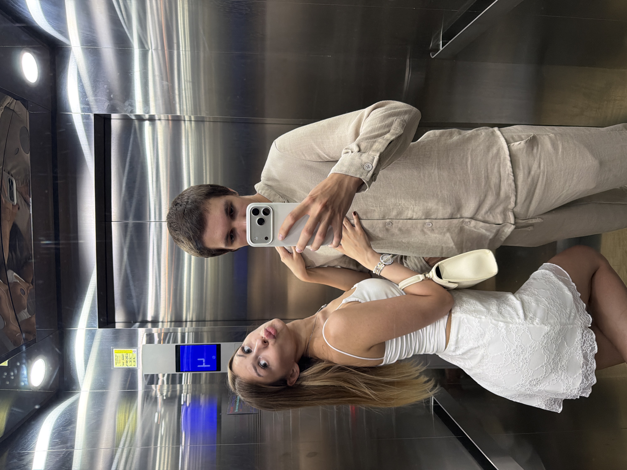 A young couple is taking a mirror selfie in an elevator. The man is on the left, wearing a light brown linen shirt. He is holding a smartphone in his right hand, which is capturing the reflection. The woman is on the right, wearing a white ribbed tank top and a white lace skirt. She is holding a white handbag on her left shoulder and making a kissy face towards the camera. Both individuals appear to be in their early to mid-twenties. The elevator has stainless steel walls that reflect their image. There is a lit panel on the wall to the left and a digital display on the right showing a "1" in a blue rectangle, indicating the elevator is on the first floor. A yellow notice is attached to the wall. The overall mood seems casual and playful. Given the location is Đà Nẵng, Vietnam, it is likely daytime or early evening.