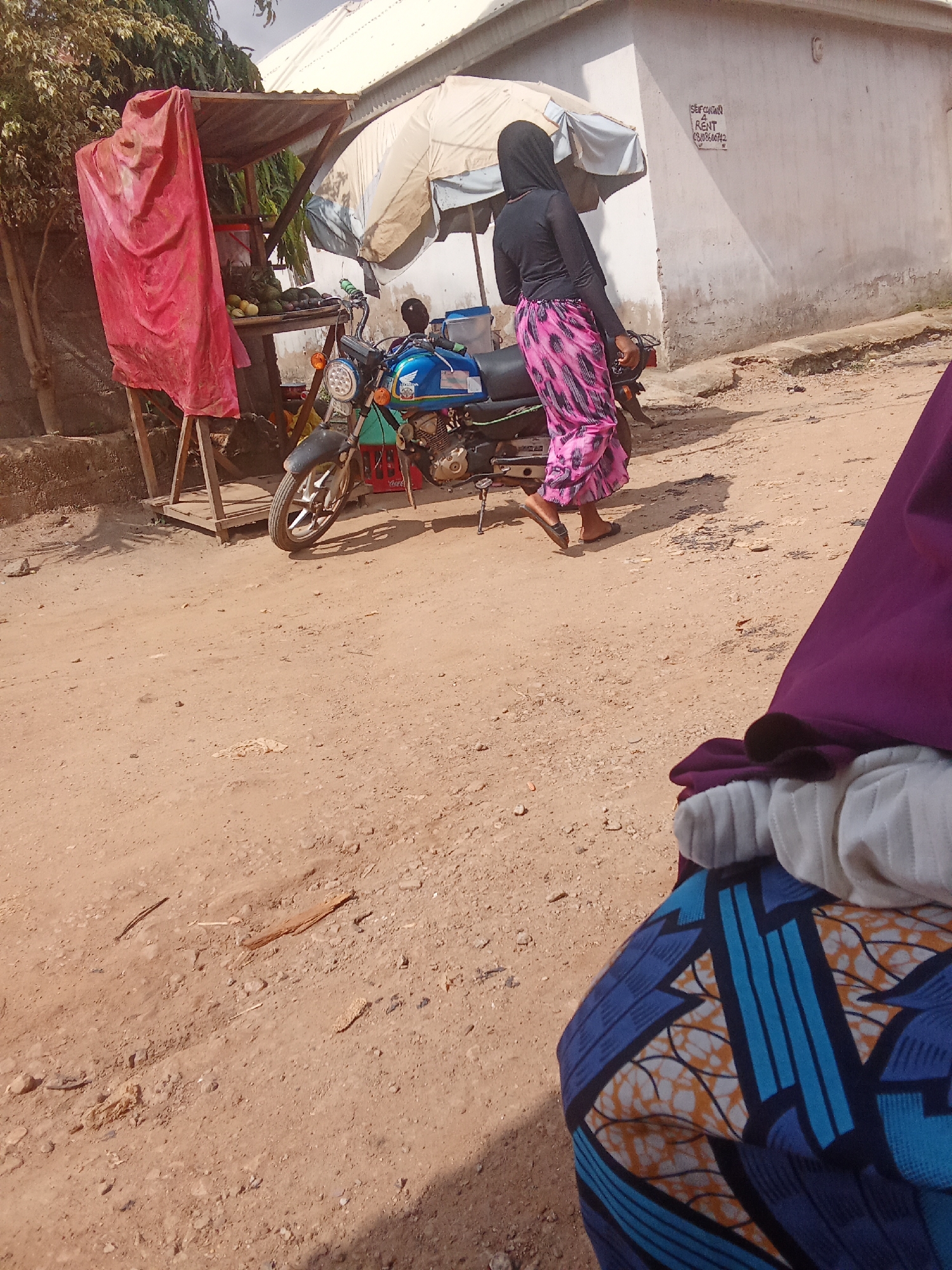 In Kagini, Nigeria, a woman wearing a black long-sleeved top and a vibrant pink and black patterned skirt walks past a roadside stall. The stall, partially covered by a red tarp and a cream-colored umbrella, displays produce, including what appear to be avocados and other fruits. A blue motorcycle is parked next to the stall. In the background, a building with a white plastered wall is visible, featuring a sign with handwritten text. The ground is dusty and uneven, characteristic of a rural or semi-urban street. The scene appears to be set during the daytime, under what seems to be a bright sky. The overall impression is one of daily life in a Nigerian community, with commerce and transportation coexisting in an open-air setting. The sign on the building is partially legible, possibly indicating a rental property or service. The woman's attire suggests modesty, common in the region. The presence of fruits indicates a local market activity.