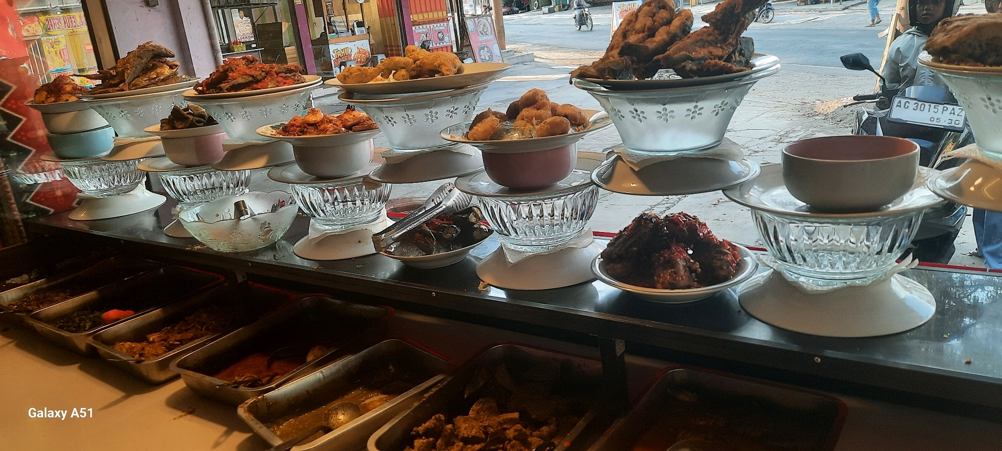 The image depicts a street-side food stall in Tangkil, Indonesia, showcasing a variety of prepared dishes. The stall features a tiered display of food, with plates of fried fish, meat dishes in sauce, and other savory items. Several glass bowls and serving dishes are used to present the food. In the background, a motorcycle with a license plate reading "AC 3015 PAZ" is visible, along with a person wearing a helmet. The overall scene suggests a bustling outdoor food vendor environment.