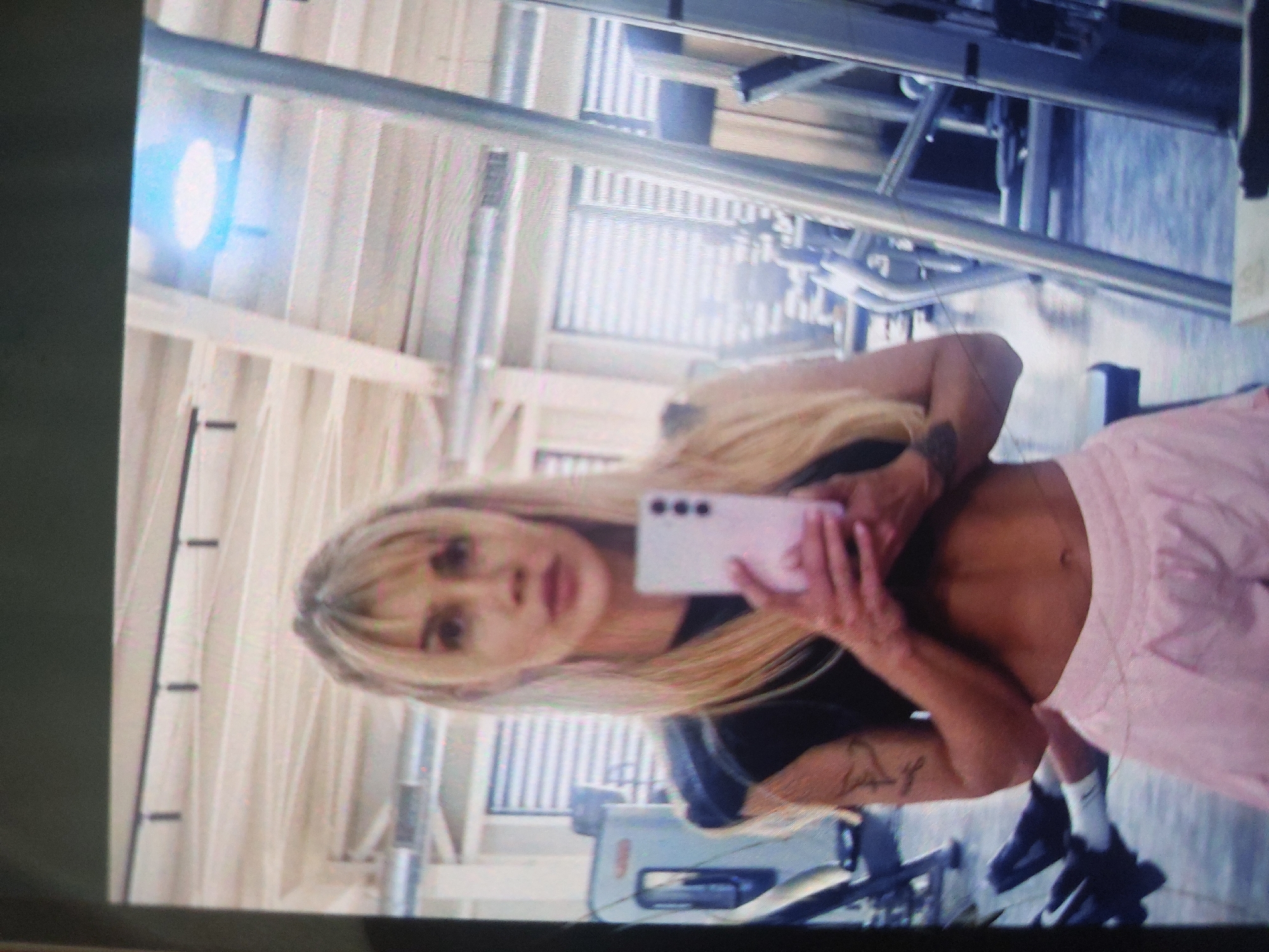 A blonde woman is taking a selfie with a pink smartphone in a gym. The woman is wearing a black shirt and pink shorts. She has tattoos on her arms. Gym equipment can be seen in the background. The location is likely in a gymnasium, but there is no definitive information in the image to confirm it is in Tsarevo, Bulgaria.