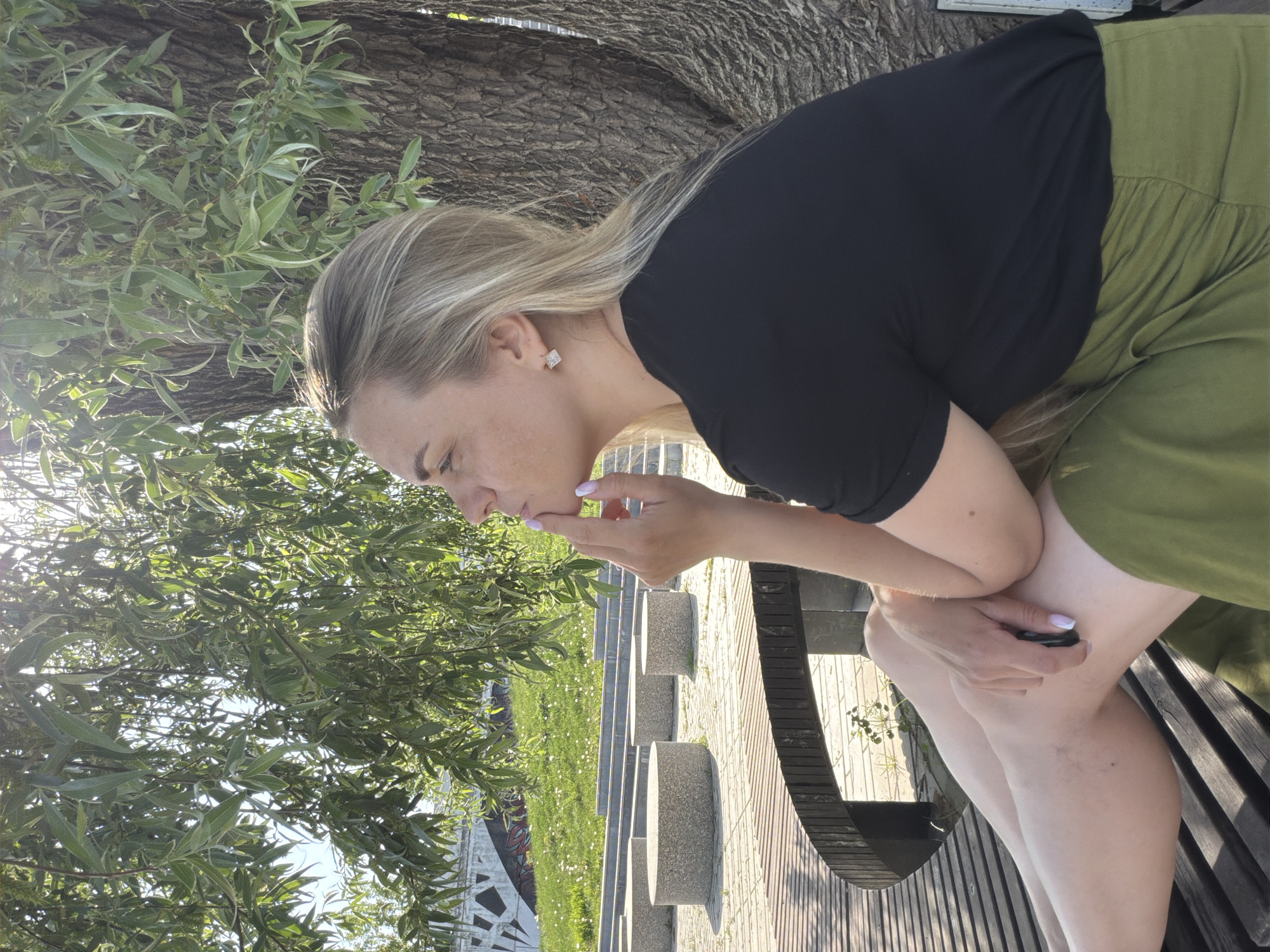 A fair-skinned woman with blonde hair is seated on a wooden park bench, possibly located in Votkinsk, Russia, as indicated by the prompt. She is dressed in a black short-sleeved top and olive-green pants or a skirt. She appears to be thoughtfully touching her chin with her right hand, her left hand resting on her lap. The woman is situated beneath a large tree with dense foliage, and the sunlight filters through the leaves. The background includes a grassy area, concrete steps or platforms, and a structure that may be a modern art installation or architectural element, potentially part of the park's design. The scene is captured during daylight hours.