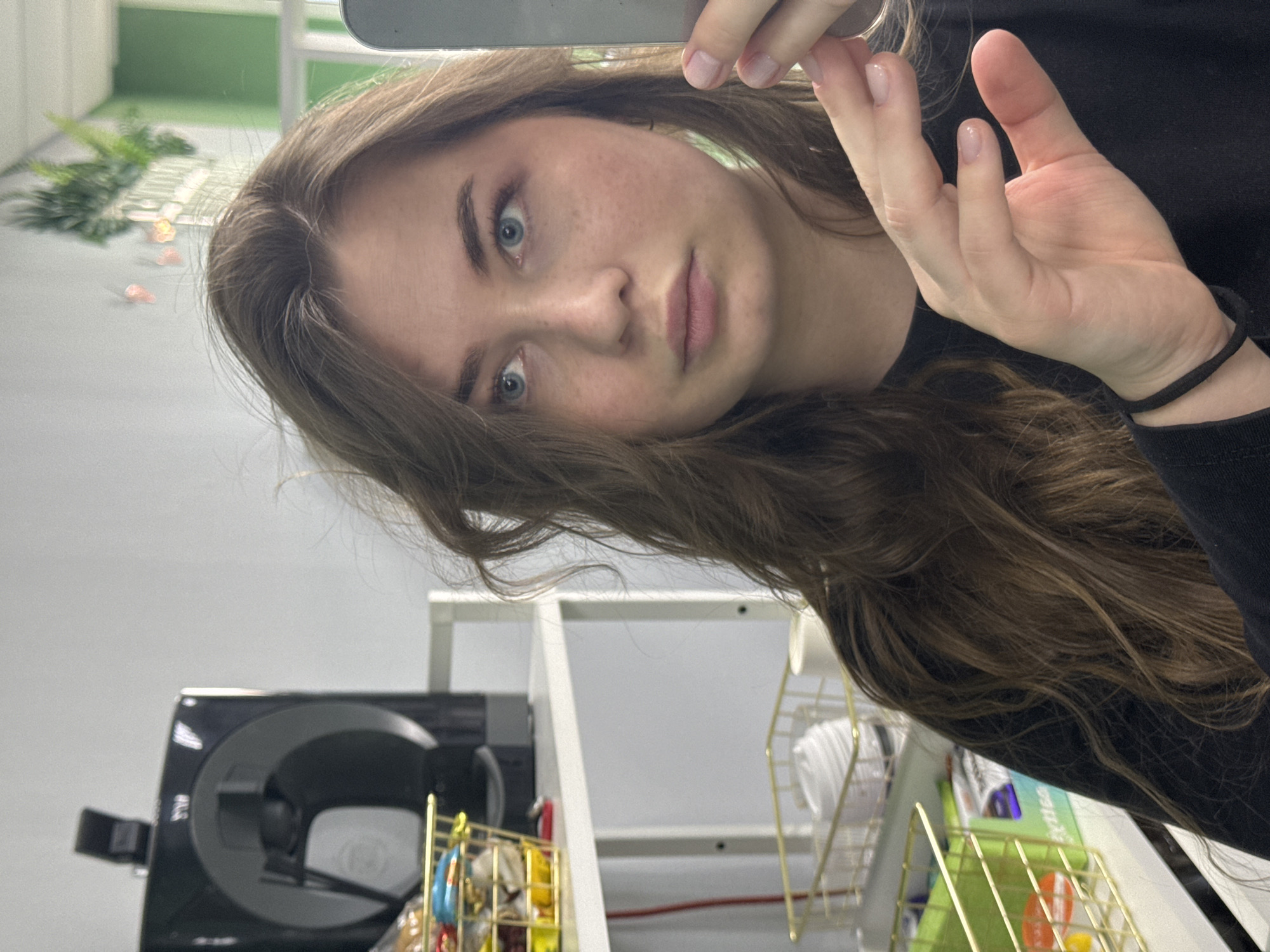A woman with fair skin, freckles, and blue eyes is taking a selfie. She has long, wavy brown hair and is wearing a black long-sleeved shirt with a black hair tie on her wrist. Her fingers are visible in the frame, possibly holding the device she's using to take the picture.
In the background, there is a cluttered shelf with various items, including a black electronic device with a circular opening, small colorful containers, a gold wire basket with white objects, and what appears to be a green box or book. The wall is white.
Based on the information provided, the location is in the city of Chaikovsky, Russia.