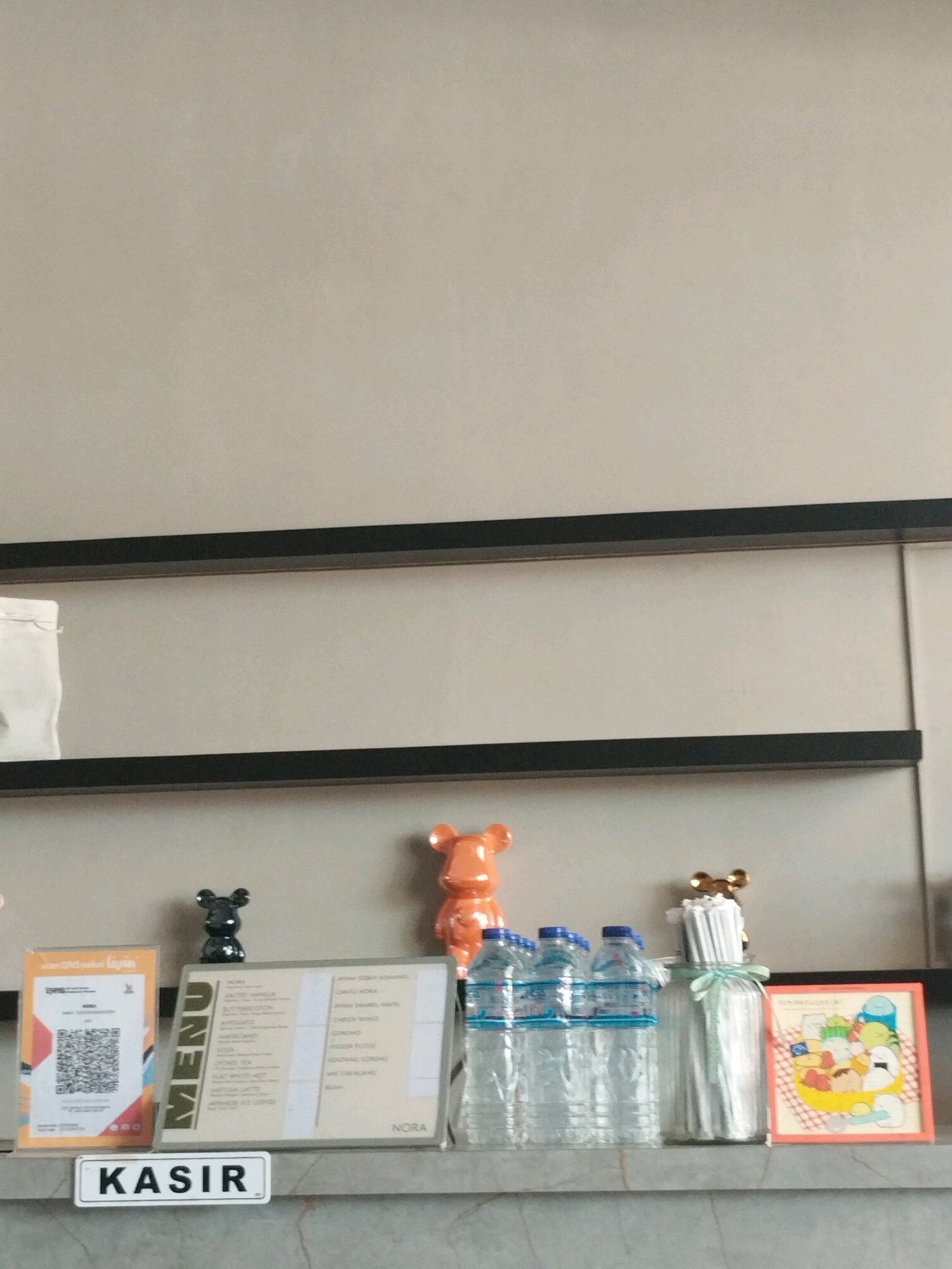 The image shows a shelf against a light beige wall. On the shelf, from left to right, are a white QR code payment stand with colorful accents, a small black bear figurine, a menu titled "MENU NORA" with a list of food and drinks, a large orange bear figurine, a row of four plastic water bottles with blue caps, a clear glass bottle filled with white straws tied with a light green ribbon and topped with a small golden bear figurine, and a framed picture with a cartoon illustration of various food items. Below these items, a white rectangular sign with "KASIR" printed in black letters is affixed to the shelf. The overall setting appears to be a counter or service area within a cafe or restaurant in Gorontalo, Indonesia, likely a point of sale indicated by the "KASIR" sign, which means "cashier" in Indonesian.