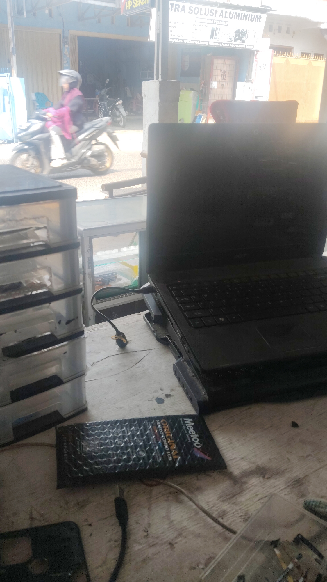 In Bandar Lampung, Indonesia, on a wooden table, a black laptop is placed on a cooling pad, connected by a cable. To the left, a stack of clear plastic drawers is visible. In the foreground, a black pouch with "Meetoo ORIGINAL" printed on it lies next to a USB cable and a smartphone component. The background shows a street scene with a person riding a motorcycle and a shop with signage indicating "HP SEKEN" and "TRA SOLUSI ALUMINIUM".