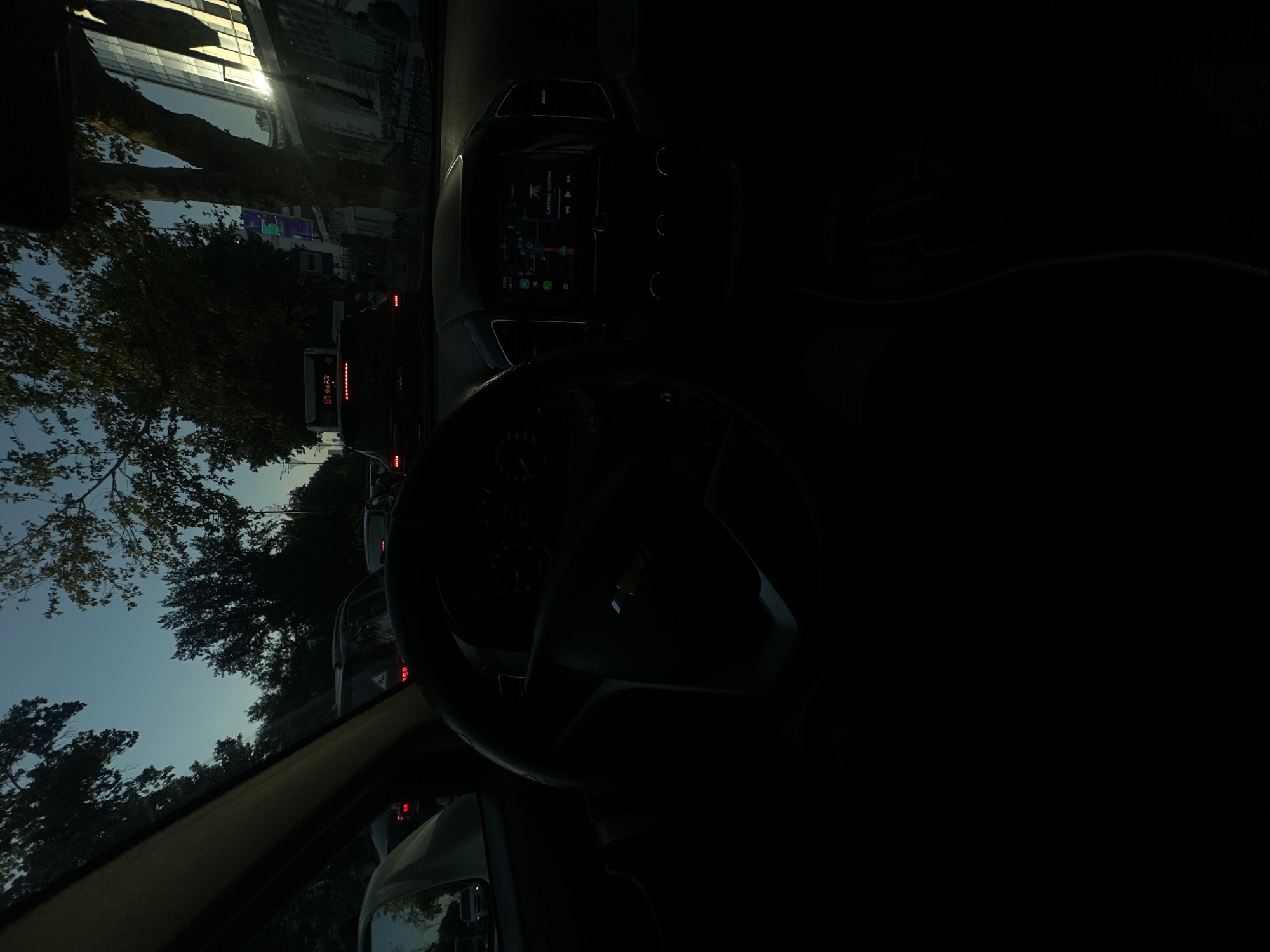 The image captures a view from inside a car, likely in Tashkent, Uzbekistan, during dusk. The perspective is from the driver's seat, with the steering wheel and dashboard prominently visible. Outside the car, a traffic jam is evident on a city street, with other vehicles ahead. Trees line the road, and a modern glass-fronted building with sunlight reflecting off its facade is visible on the right. The sky is a twilight blue, indicating the transition from day to night. The dashboard display shows a navigation map and what appears to be media playback controls, suggesting the car's infotainment system is active. There are no discernible individuals in the frame, other than potentially reflections.