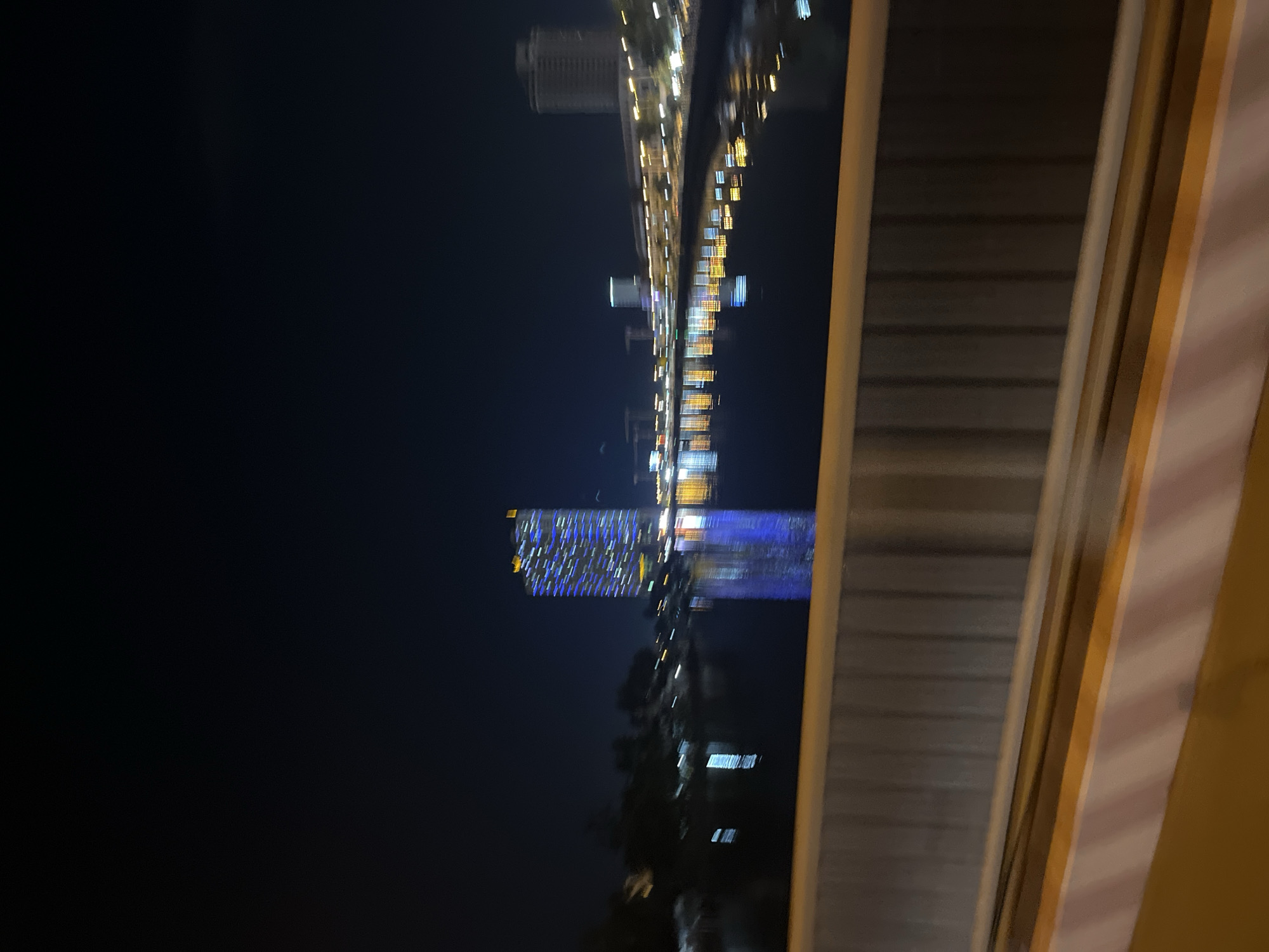 The image shows a nighttime view from a window in the Nha Trang Ward, Vietnam. The scene is dark, with a skyline featuring illuminated buildings reflected in water. There are trees in the foreground along the water. A window frame is visible on the right side of the image.