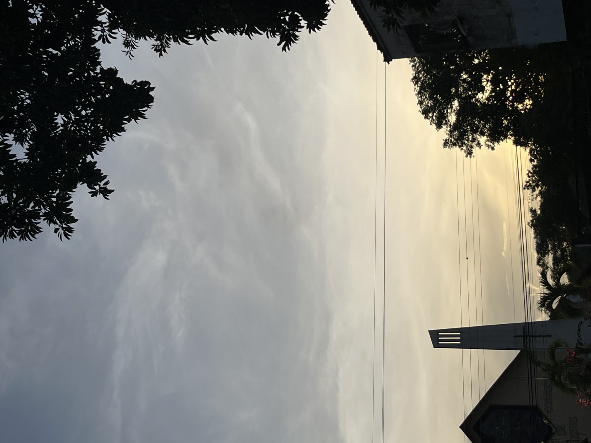 The image shows a view looking upward, framed by trees on the left and upper right, and buildings on the upper right and lower right. Power lines traverse the sky. The sky transitions from a blue-grey on the left to a yellowish hue on the right. A church building is visible in the lower right, featuring a distinctive tower with three horizontal openings. The image was taken in Tinaquillo, Venezuela.