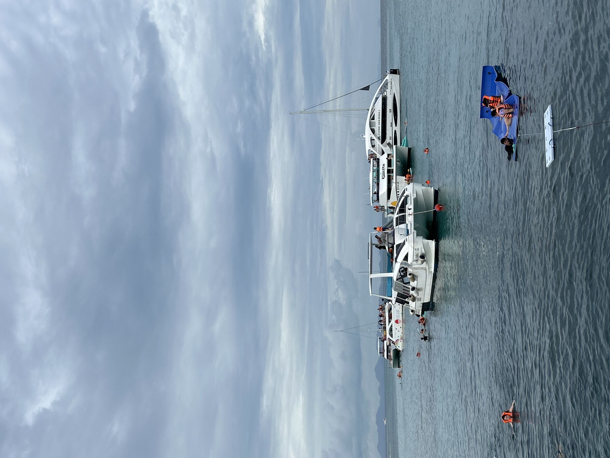 The image depicts a cloudy day at sea in Rawai, Thailand, with several boats and people engaged in water activities. Multiple white catamarans are anchored in the calm, blue water. One catamaran, labeled "FLYING DOLPHINS 1," is prominently featured, as is another with a colorful design and the label "TIP TOP." There are many people on the decks of these boats, some appearing to be tourists. In the water, several individuals are swimming or snorkeling, some wearing orange life vests. In the foreground, a blue inflatable water slide-like structure is floating, with two people, one wearing a life vest, on it. Further in the foreground, a person in an orange life vest is swimming. In the distance, a faint outline of islands can be seen against the cloudy sky. The overall scene conveys a sense of leisure and recreation at a coastal location.