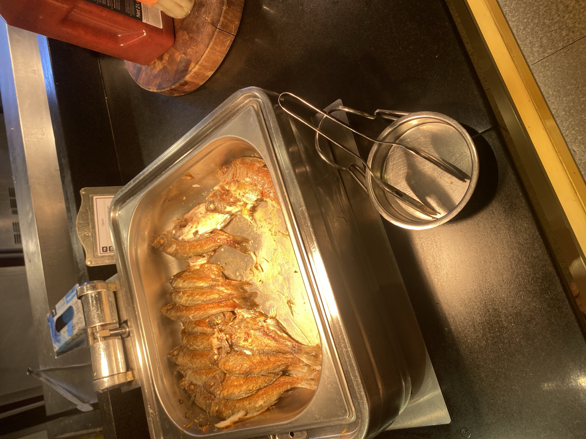 The image displays a buffet-style food serving area. In the foreground, a metal serving tray holds a large quantity of whole, fried fish, appearing to be a popular dish. Adjacent to the tray, a set of metal tongs rests in a small metal dish, ready for use. The background shows a stainless steel warming unit, typical of buffet setups. To the right, a large red bottle, possibly of sauce, stands on a wooden cutting board. The overall environment appears to be an indoor establishment, such as a restaurant or hotel dining area. The image does not contain any information indicating a specific location in the city of Aksu, Turkey.