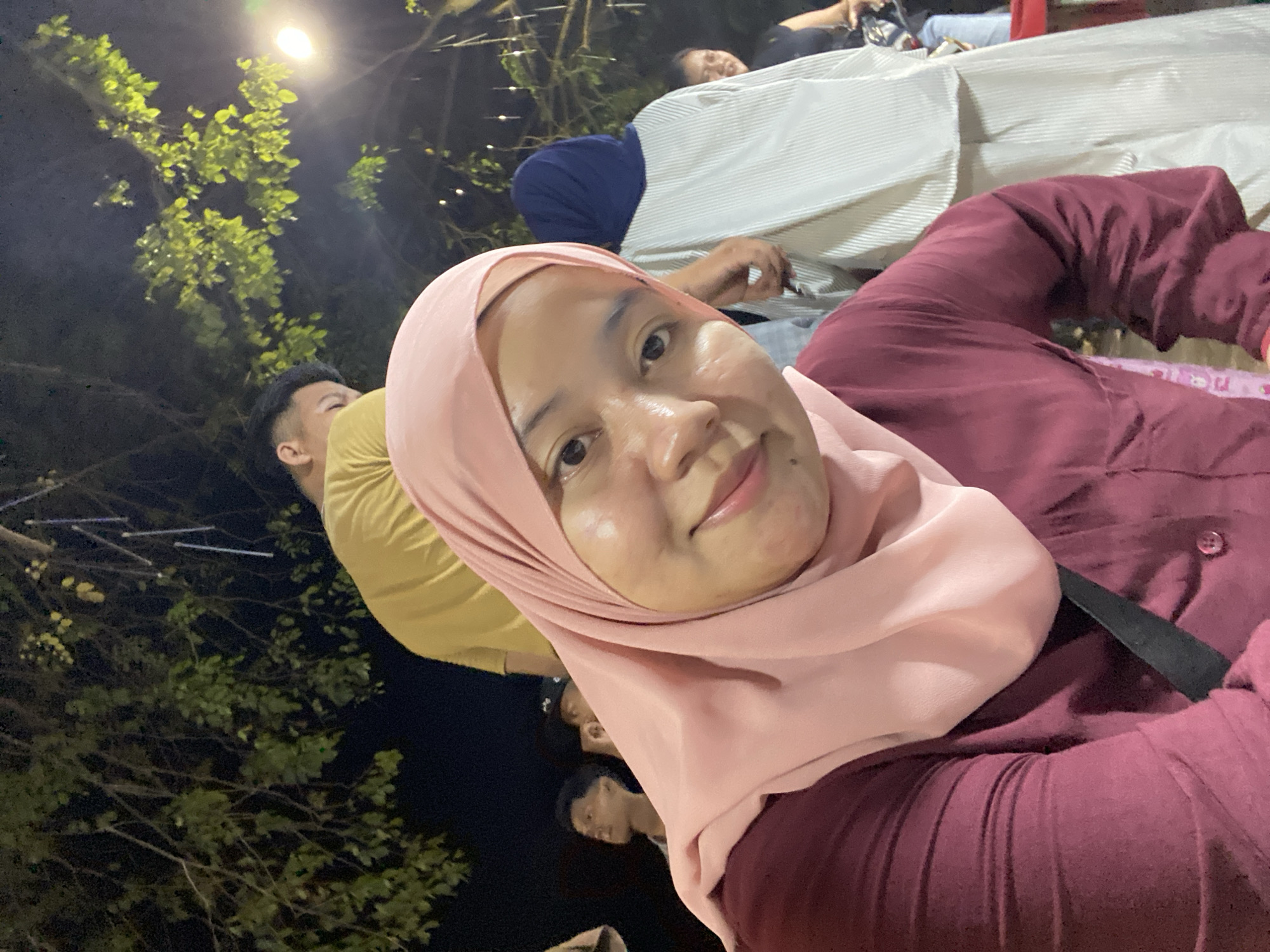 In this frontal, medium shot, a woman wearing a pink hijab and a maroon shirt is the primary subject, looking directly at the camera with a gentle smile. Her right shoulder is visible, adorned with a black strap across her chest. Behind her, the scene unfolds at night, outdoors, under a canopy of lush green foliage illuminated by string lights and a bright overhead lamp.
Several other individuals are partially visible. To her left and slightly behind, a man in a yellow t-shirt is partially obscured, his head tilted upwards. Further back, two young men are visible; one in a dark shirt looking to the left, and another in a dark cap looking upwards and to the left. To the right of the main subject, a person in a white outfit with vertical pleats and a blue hijab stands facing away from the camera. Another person is visible further to the right, partially obscured by the white outfit, wearing a dark top. A red object, possibly a table or bin, is visible at the bottom right edge of the frame. The overall ambiance suggests a gathering or event taking place outdoors at night, possibly in a park or a market area within Banjarmasin, Indonesia, given the presence of tropical foliage and the festive lighting.