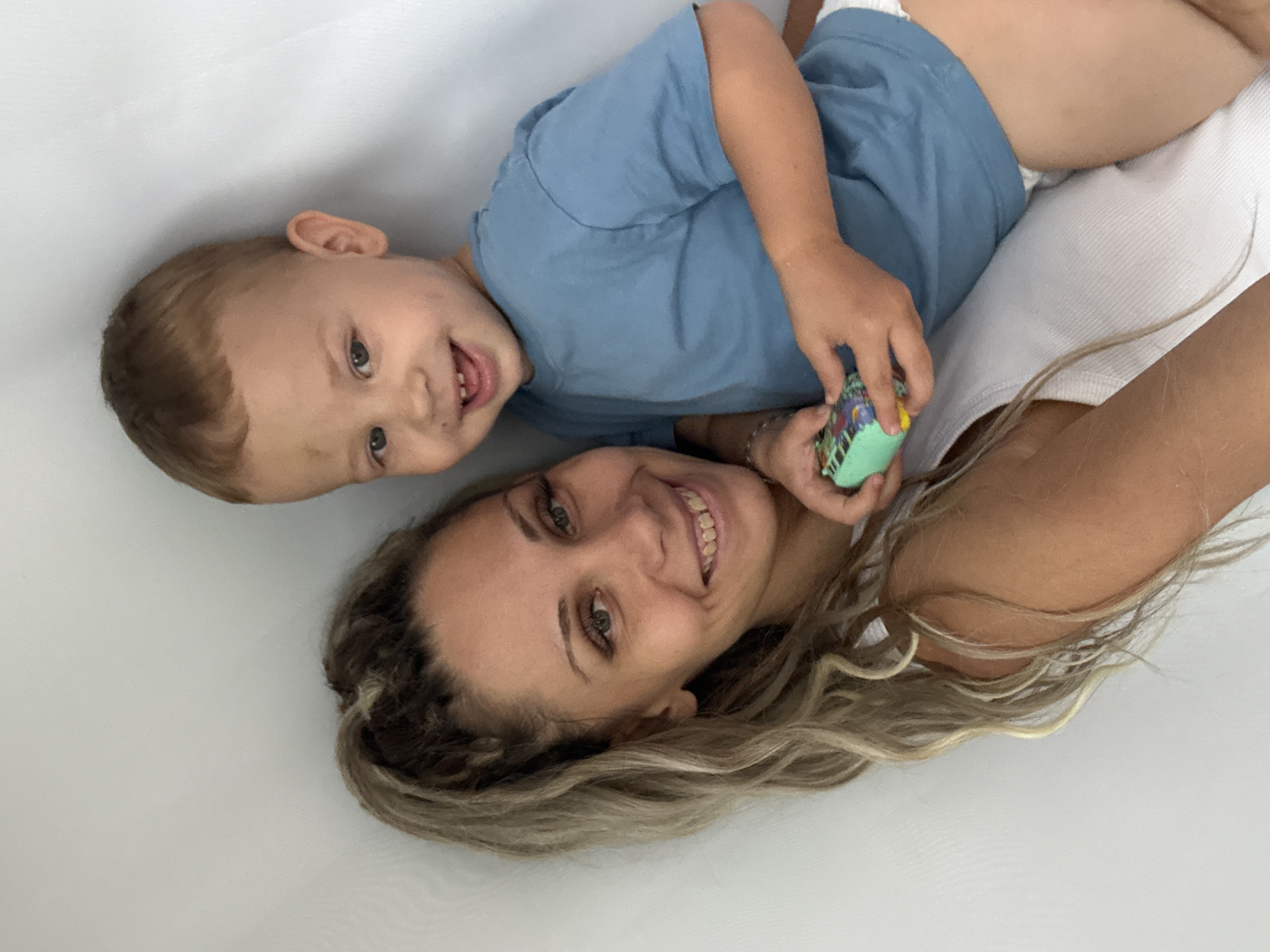 In Rusanovo, Russia, a woman with long, blonde, braided hair pulled into a high ponytail is leaning against a plain white wall. She is wearing a white tank top and smiles warmly at the camera. A young boy, appearing to be around three years old, stands in front of her, leaning his body against her. He has short brown hair and is wearing a blue t-shirt and a diaper. He is also smiling and holding a small, colorful toy in his right hand. The boy has a smudge of dirt on his forehead. The overall impression is a candid, affectionate moment between a mother and child.