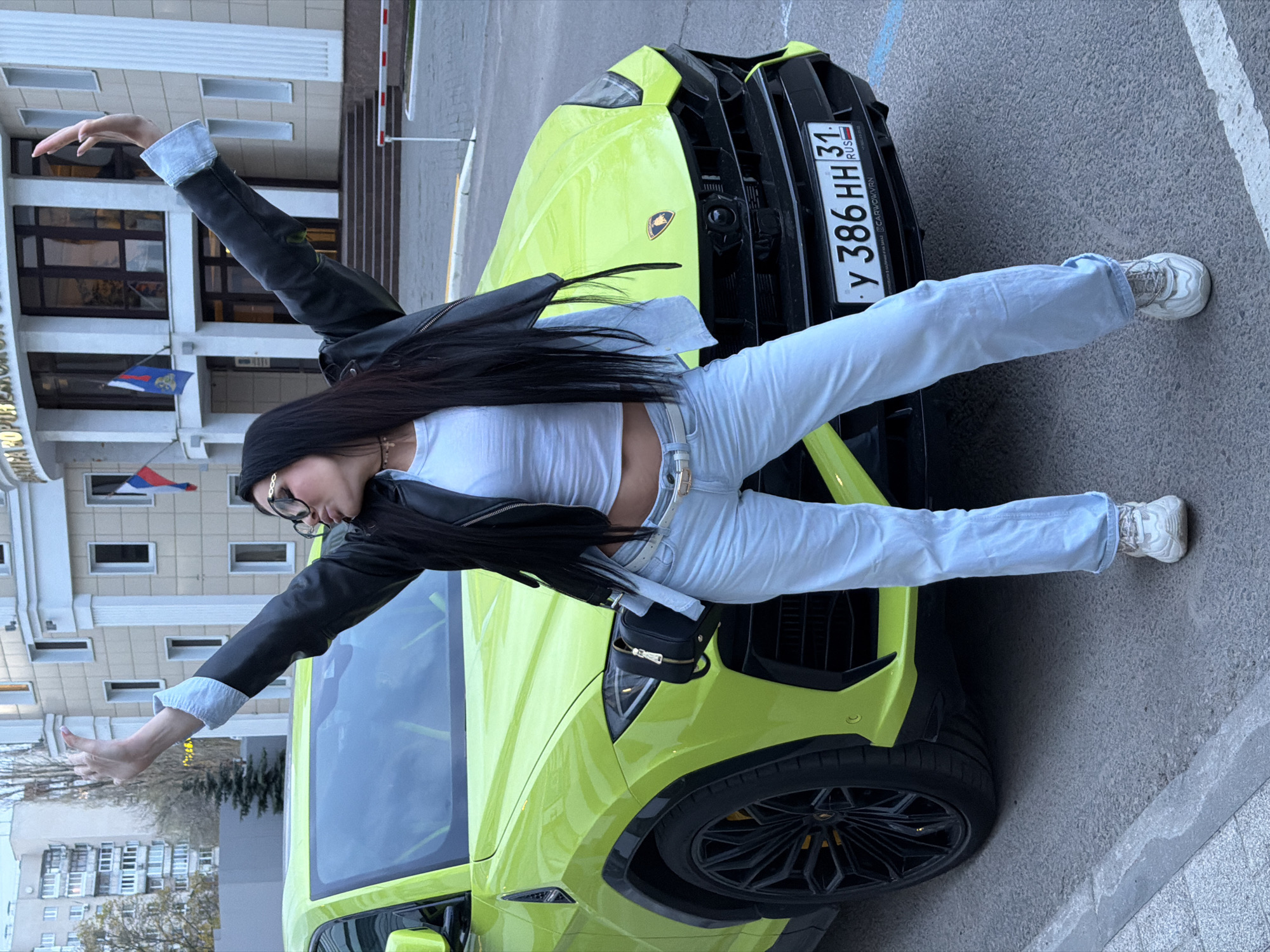A young woman is sprawled across the hood of a lime green Lamborghini Urus. She is wearing a white crop top, light blue jeans, a black leather jacket, and white sneakers. She has dark hair and is wearing glasses. Her arms are outstretched. The car's license plate reads "У 386 HH 31 RUS". The background shows a building with a Russian flag visible on its facade. The location is likely Voronezh, Russia.