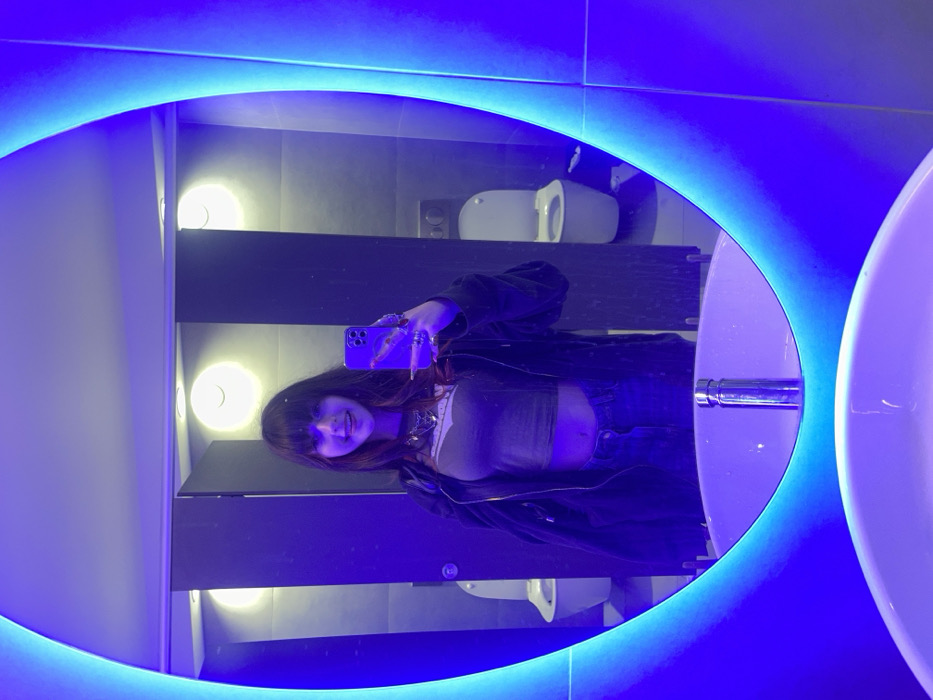 A young woman takes a mirror selfie in a public restroom. She wears a black jacket, jeans, and a light top. Her long nails are visible as she holds a purple phone. Reflected in the illuminated oval mirror is a toilet, a row of lights, gray tile, and sinks. Blue light dominates the scene, emanating from the mirror's frame.