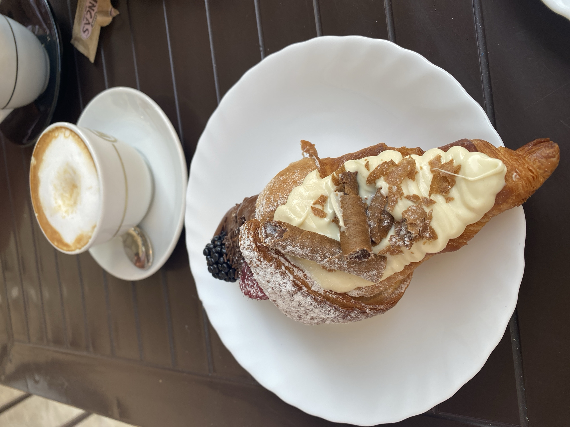 An overhead shot captures a breakfast scene on a dark brown, slatted outdoor table in Chiavari, Italy. A white plate holds a croissant filled with cream and topped with chocolate, raspberries, blackberries, and wafer cookies. To the right, a white coffee cup with a saucer holds a cappuccino, and a packet of sugar sits nearby.