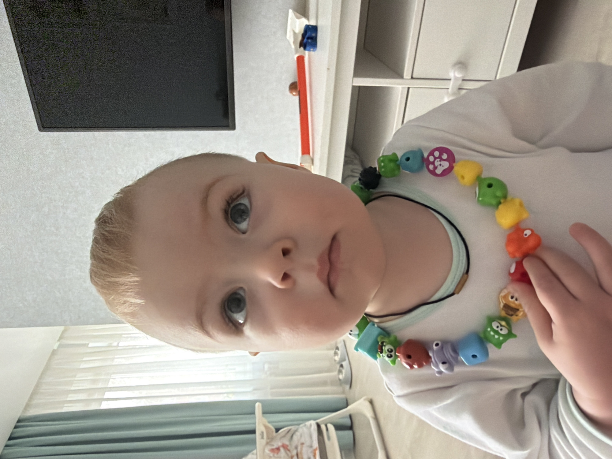 A young child with light blonde hair and light eyes is centered in the frame, wearing a white shirt and a colorful beaded necklace. The child is looking directly at the camera. A flat-screen television is mounted on the wall in the upper left corner. A white dresser or shelf is visible behind the child in the upper right corner. A window with blue curtains and a baby swing are in the background. The location is likely indoors in Votkinsk, Russia.