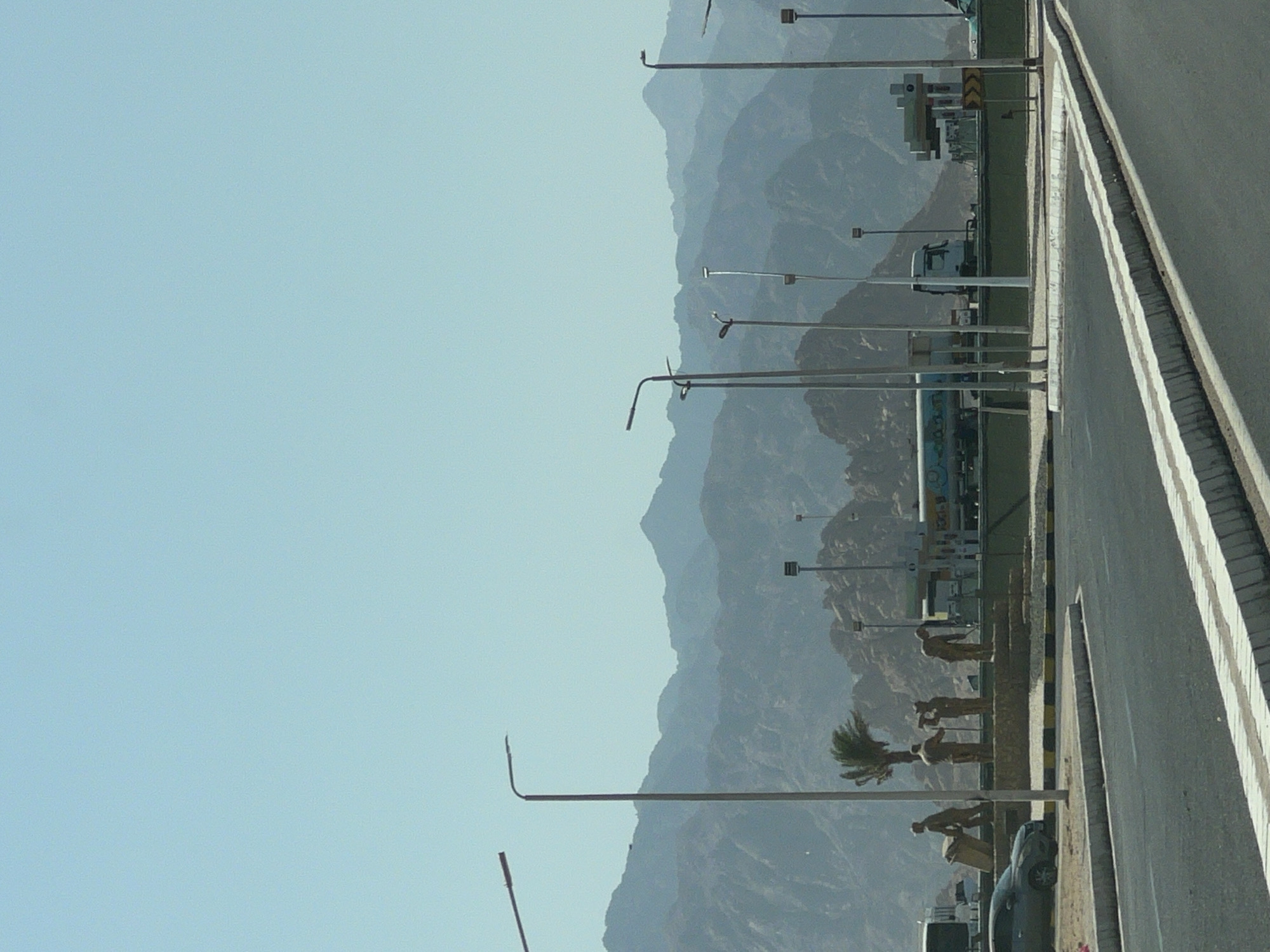 The image captures a roadside scene in Sharm El Sheikh, Egypt, with a backdrop of layered, muted blue mountains under a clear, pale blue sky. In the foreground, a paved road curves into the frame, bordered by white and black striped concrete barriers. To the left, a dark-colored sedan is parked. Beyond the road, a series of statues depicting figures in work poses stand on a raised platform, accompanied by a single palm tree. Behind the statues, a gas station with pumps and a tanker truck are visible, along with several other vehicles. Tall, slender lampposts are scattered throughout the scene, some with curved tops. The overall impression is of a functional, potentially arid, roadside environment in a mountainous region.