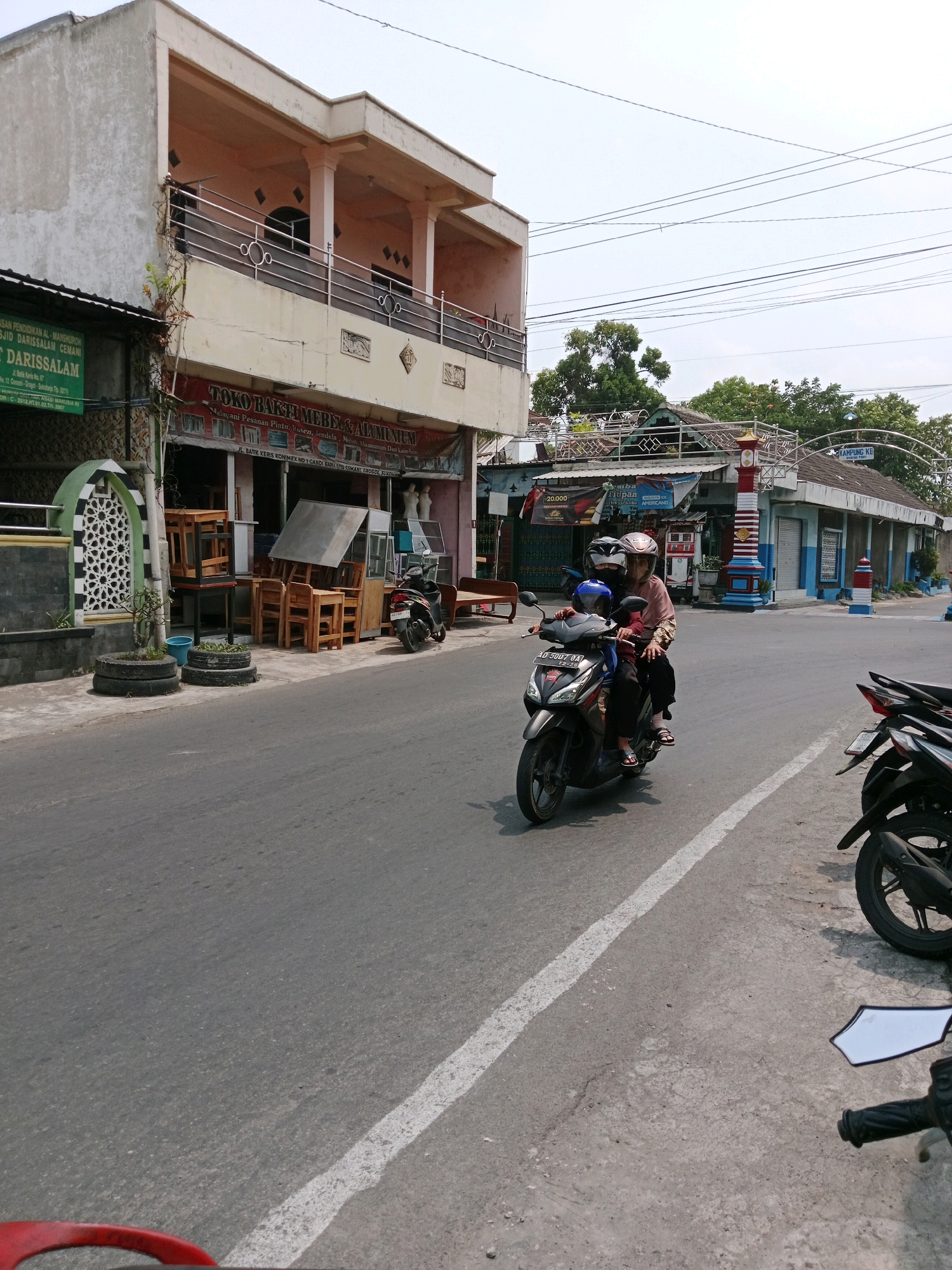 In Cemani, Indonesia, on a bright, clear day, a street scene unfolds. A two-story building with a pale facade and a railing balcony dominates the left side of the frame. Below, a shop with a red awning displays furniture for sale, including wooden chairs and tables. To the left of the shop, a green sign reads "MASJID AL-MANSHUROH" and "DARISSALAM". On the sidewalk, potted plants in repurposed tires add a touch of greenery. A street with a white line marking the lane stretches towards the horizon. In the foreground, a black scooter carries two individuals, a woman in a pink shirt and headscarf and a child in blue, both wearing helmets. Another parked black scooter is visible on the right side of the street. The environment appears to be a commercial area with various shops and buildings lining the street.