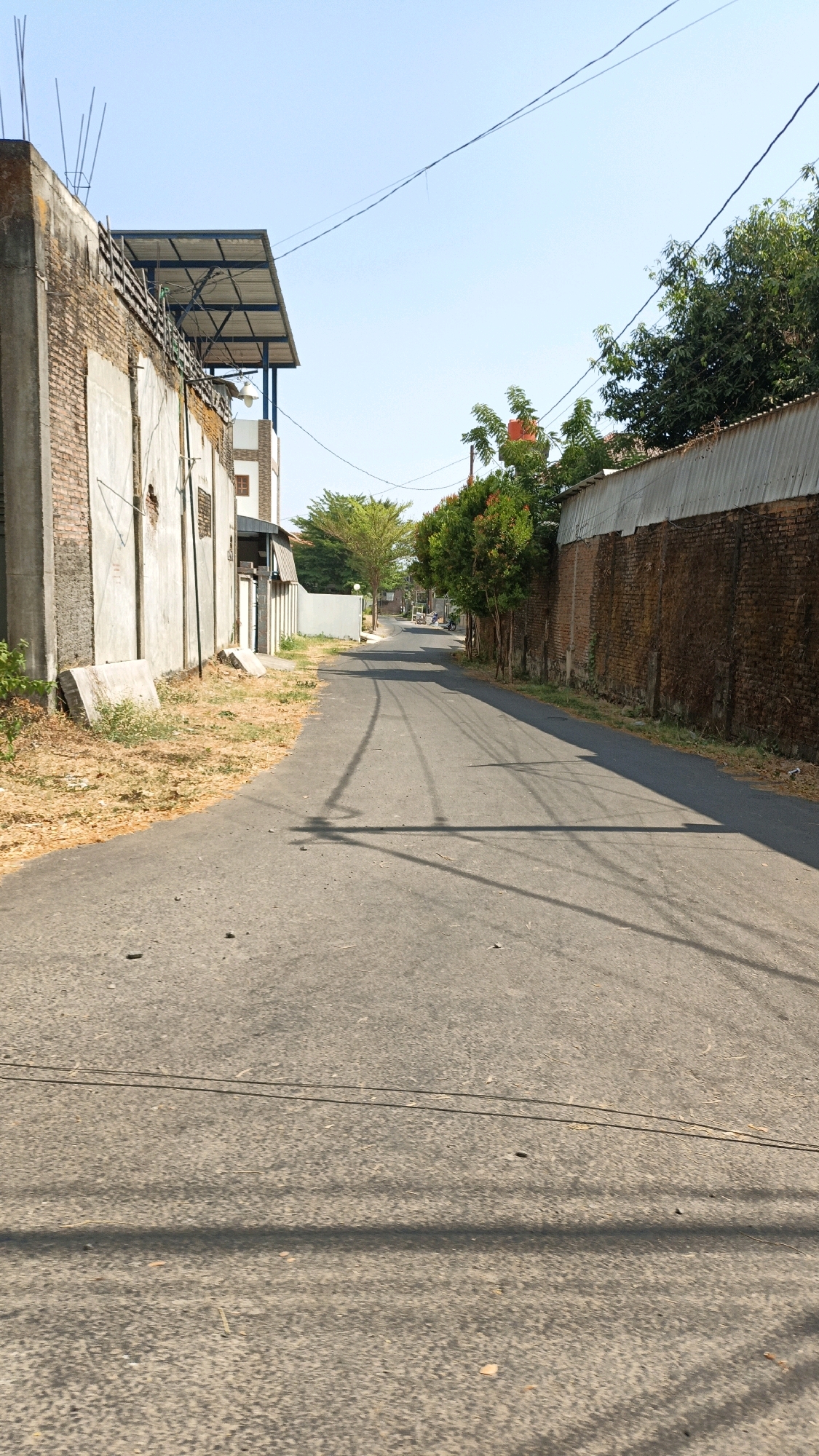 The image shows a narrow, paved road in the city of Cikal, Indonesia, bordered by buildings and vegetation. The road curves slightly to the right as it recedes into the distance. On the left side of the road, there is a brick building with a concrete facade and a metal roof structure. Dry grass and some concrete blocks are visible on the side of the road next to this building. On the right side of the road, a brick wall topped with corrugated metal runs along the property line, with trees and foliage growing behind it. Further down the road, more buildings and trees are visible, and in the distance, two people on a motorcycle can be seen. The sky is clear and blue, indicating it is daytime. The scene appears to be a residential or mixed-use area in a tropical climate.