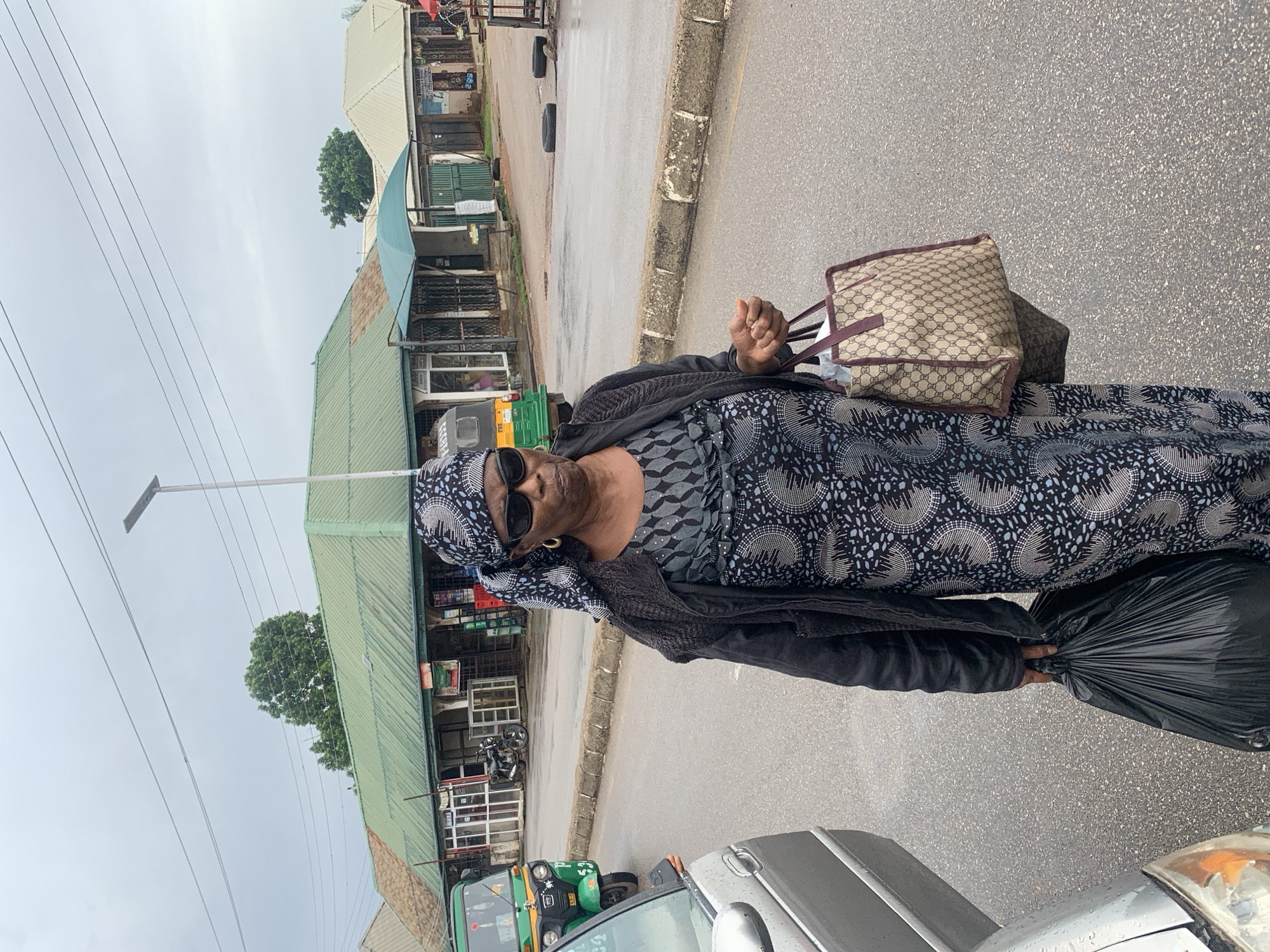 The image shows an overcast day in Jos, Nigeria. A woman in a headscarf and a patterned dress is walking on a wet road, carrying a black trash bag in her left hand and a designer tote bag in her right. In the background, there are several shops with green roofs, some with awnings. A green auto-rickshaw and a silver car are also visible on the road. The overall scene suggests a market or commercial area in the city.