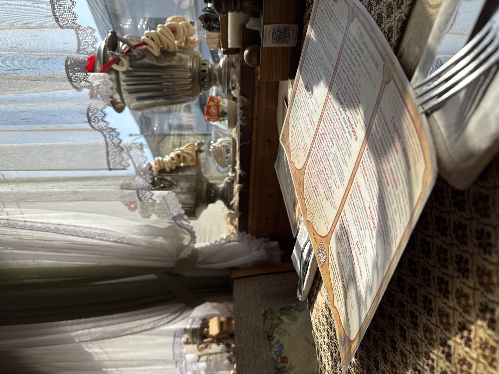 Here is a description of the image:
The image depicts a restaurant or cafe setting. A menu is prominently displayed, detailing various dishes, including soups, dumplings, and main courses, with prices listed in Russian currency. In the background, there's a samovar, a traditional Russian tea urn, indicating the establishment's cultural or historical theme. Light streams in through a window covered with a lace-trimmed curtain. The table is covered with a patterned cloth. The image evokes a sense of traditional Russian dining. The mention of "Ростовская" in the menu suggests the restaurant or establishment might have a specific regional culinary focus.