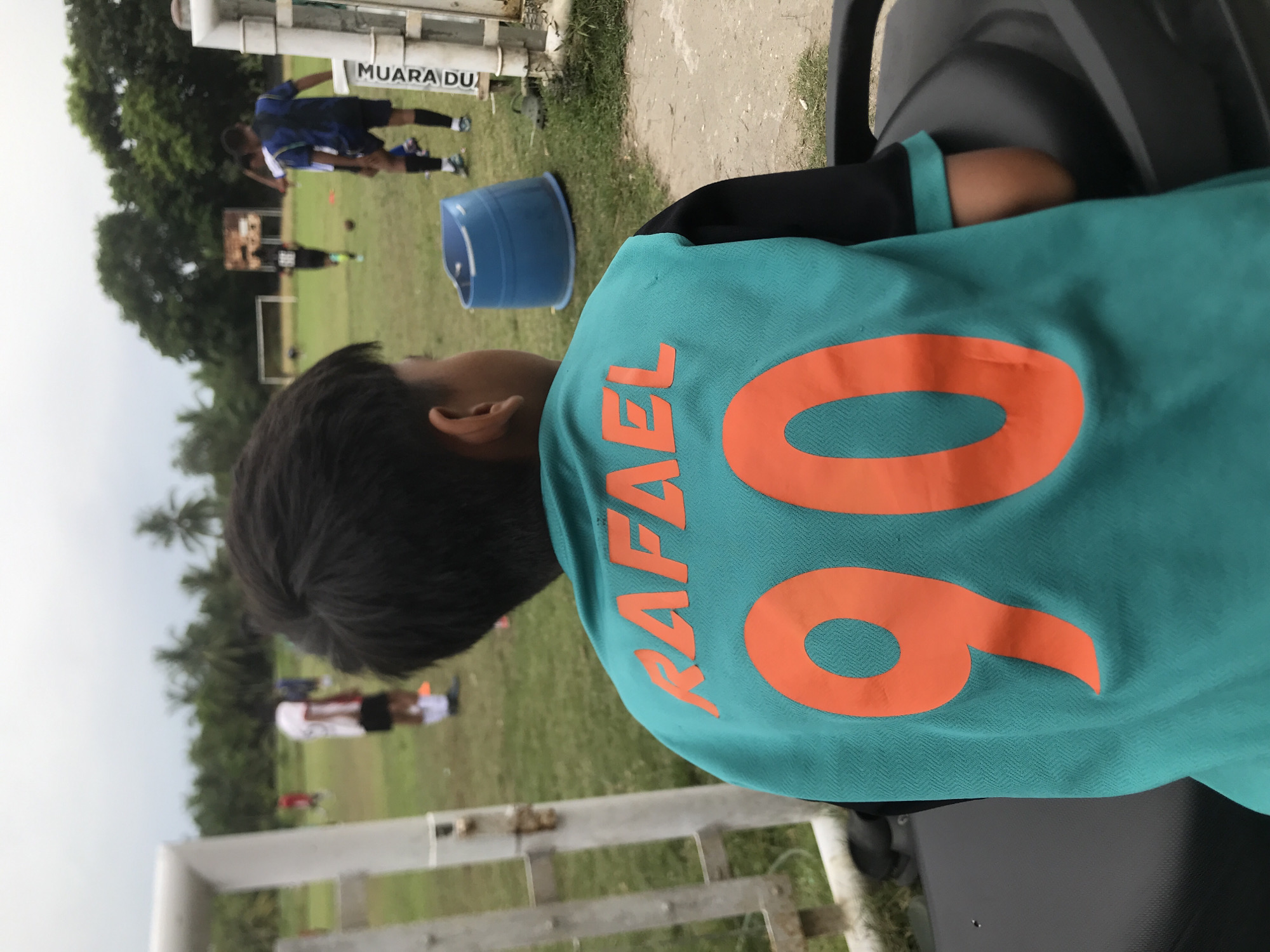 The image shows a child wearing a teal soccer jersey with the name "RAFAEL" and the number "06" in orange lettering. The child is sitting, possibly on a bench, facing away from the camera and towards a soccer field where other people are practicing. A blue bucket sits on the grass between the child and the soccer players. The location appears to be outdoors, with trees visible in the background under a cloudy sky. A sign that reads "MUARA DU" is partially visible on a fence near the soccer field. The location of the image is likely in Palohlada, Indonesia.