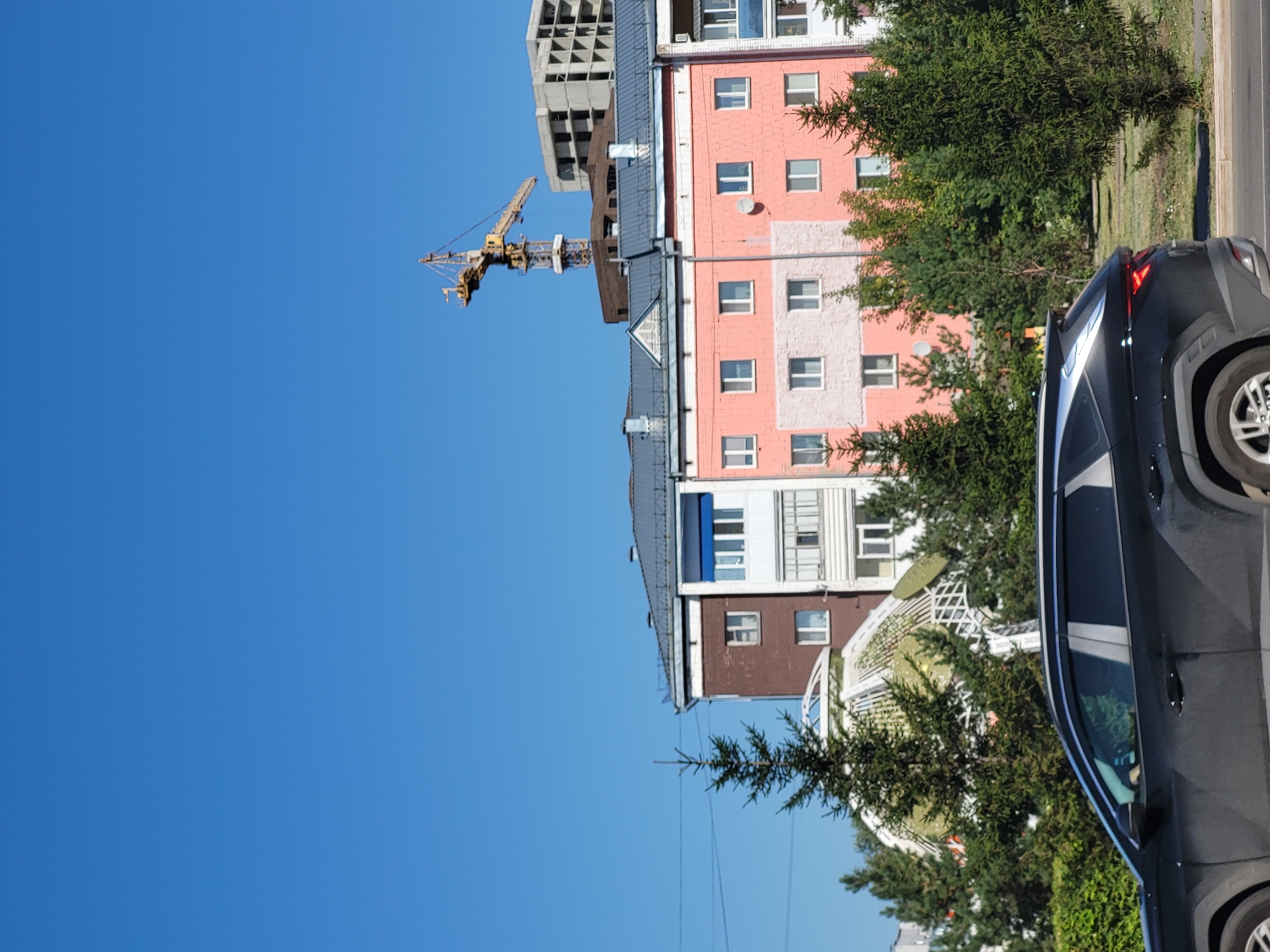 The image depicts a street scene in Petropavl, Kazakhstan, under a clear blue sky. In the foreground, a dark-colored SUV is driving on a paved road. Behind the vehicle, a row of lush green coniferous trees lines the street, some of which partially obscure the buildings beyond.
The buildings in the background include a multi-story apartment building with a pink facade and white window frames, another building with a dark brown and white facade, and a partially constructed concrete building. A prominent yellow construction crane is visible above the buildings, indicating ongoing construction in the area. An unusual, dome-like white structure, possibly part of a greenhouse or conservatory, is also visible behind some of the trees. The overall impression is a snapshot of urban life in Petropavl, combining residential areas with ongoing development.