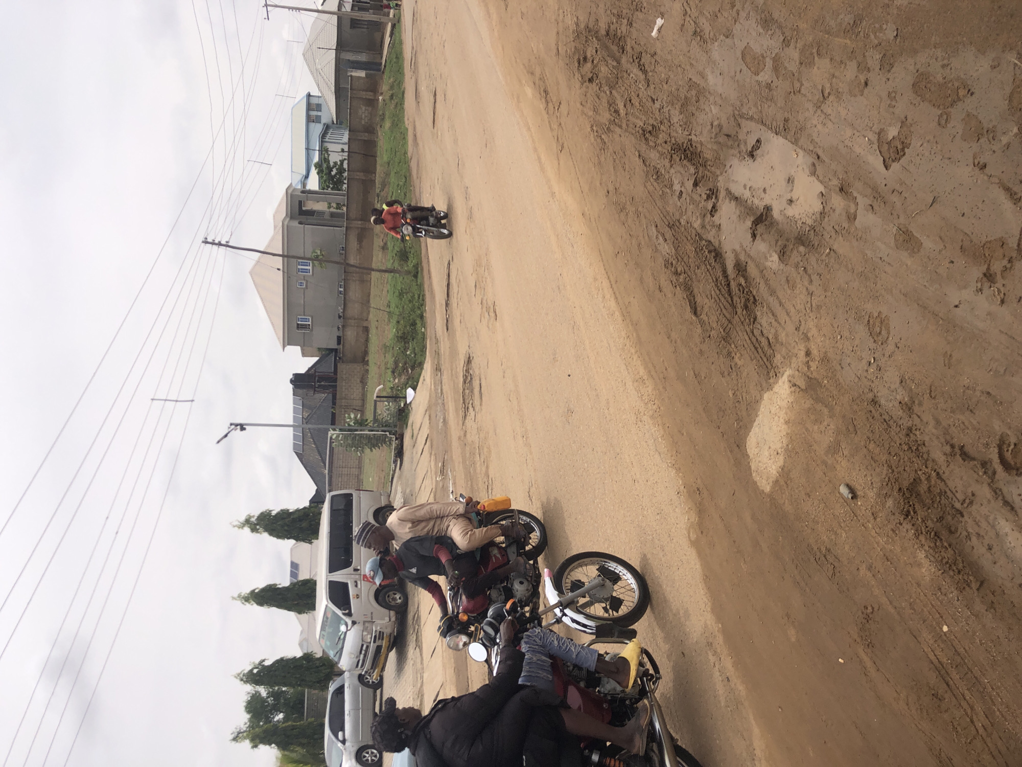 In Hanwa, Nigeria, the image captures a street scene with a dirt road. Two motorcycles are visible; one is parked with multiple riders, while the other is further down the road. Adjacent to the road is a row of parked vehicles, including a van and cars. Buildings are visible in the background, interspersed with trees and utility poles. The sky is overcast.