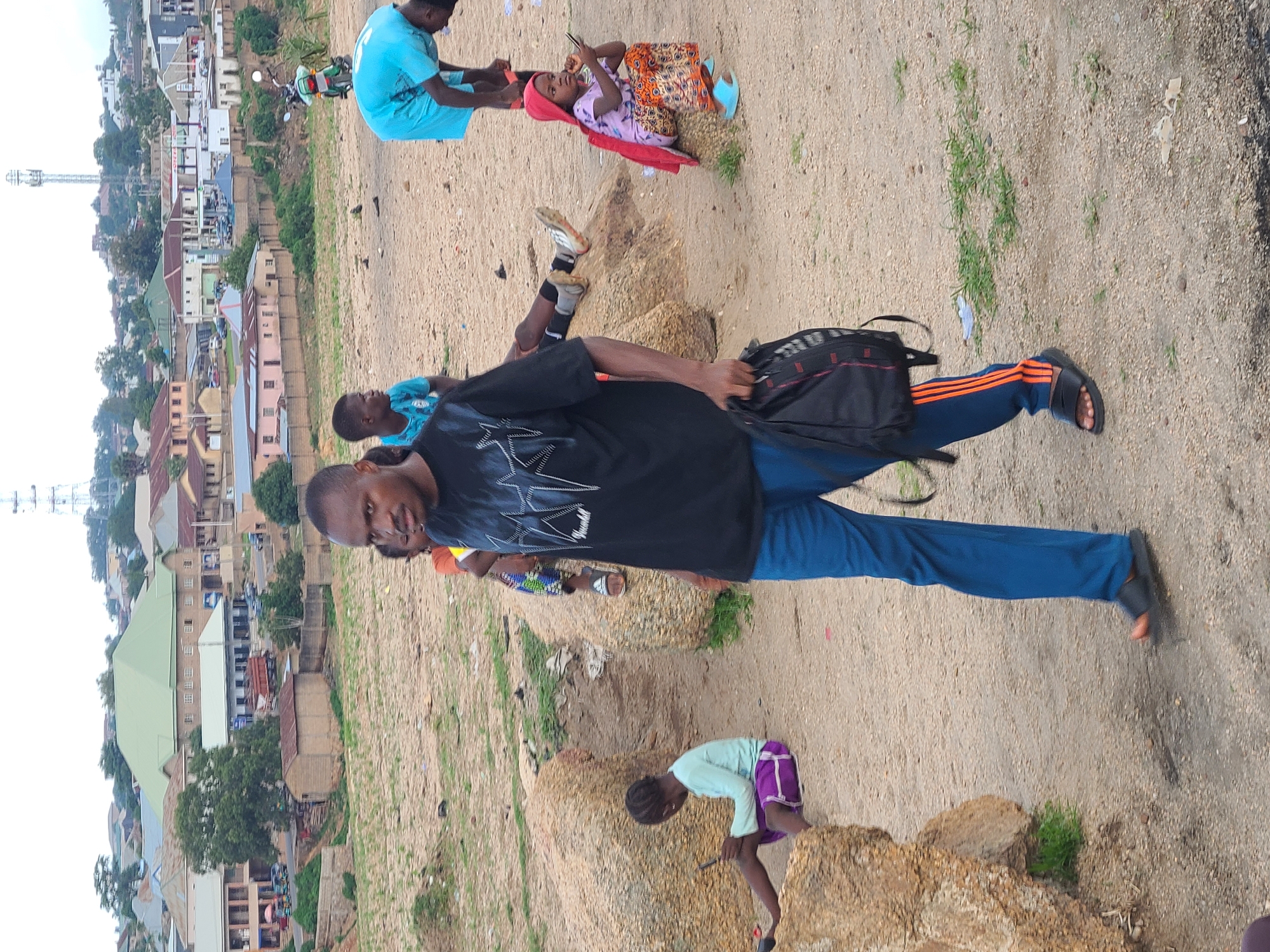 In Jos, Nigeria, a man in a black t-shirt and blue pants walks towards the camera, carrying a black backpack. He is in the foreground, with other people and a cityscape in the background. A young girl in a light blue long-sleeved shirt and purple shorts is seated on rocks to the left, looking at a phone. To the right, a person in a light blue shirt is bending over, and a young girl in a pink outfit and red head covering is sitting on a rock, also looking at a phone. In the middle ground, other individuals are visible, including a person lying down on rocks. The background shows a dense urban area with buildings of various sizes and colors, a prominent green-roofed structure, and a radio tower in the distance. The ground is sandy with scattered rocks and sparse vegetation.