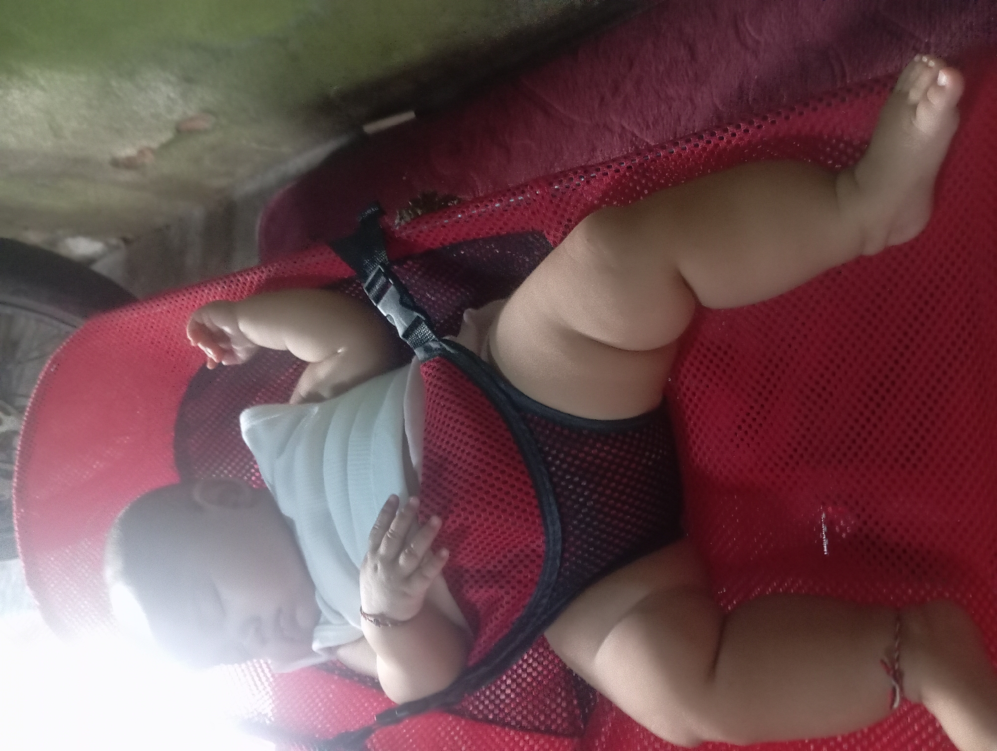 A baby is sleeping in a red bouncer seat. The baby is wearing a white tank top and dark shorts. The baby has a bracelet on its left wrist and anklets on both ankles. The bouncer seat is placed on a red fabric surface. The background appears to be an outdoor setting with a green wall and some debris on the ground. The lighting suggests it is daytime. The location is specified as Rejosari, Indonesia.