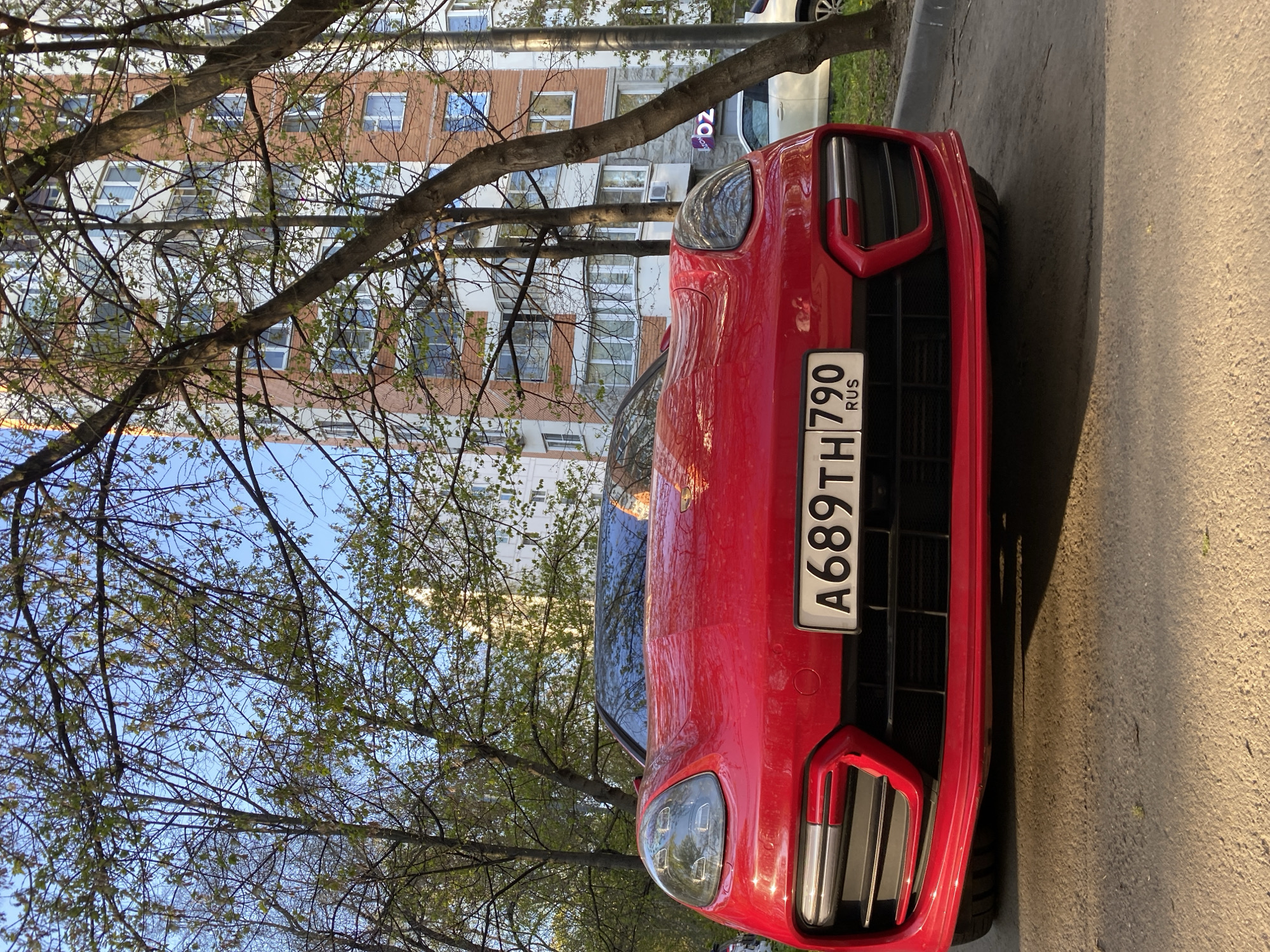 A red car is parked on the side of a road near an apartment building with trees in Moscow, Russia. The car's license plate number is A689TH790. The car is parked on asphalt. The building is red brick with white window frames.