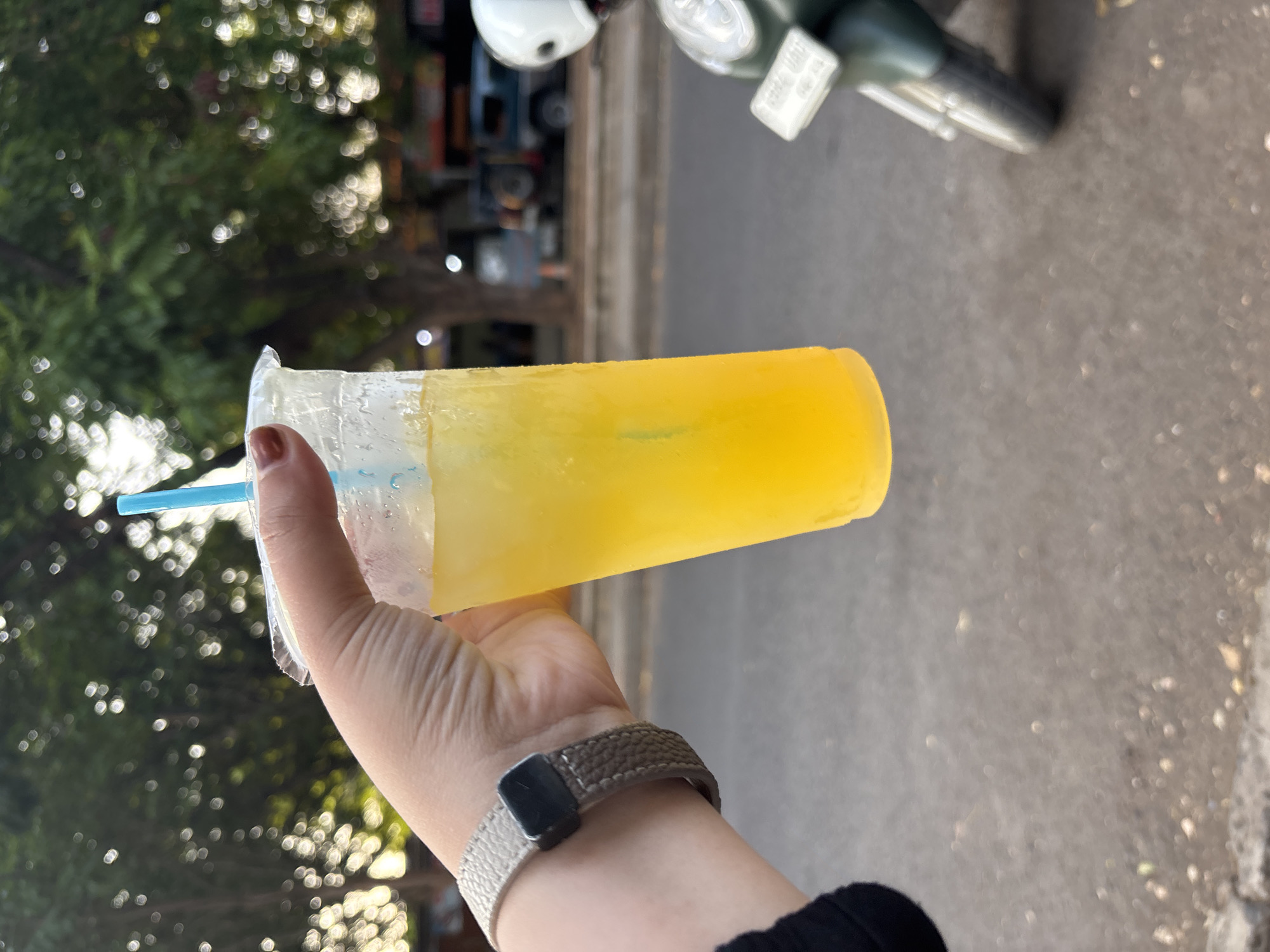 A hand with brown nail polish holds a clear plastic cup filled with a bright yellow drink and ice, topped with a blue straw. The cup is slightly fogged, indicating it contains a cold beverage. The hand wears a textured grey watch band. The background is blurred, showing lush green trees with dappled sunlight, a paved road, and hints of buildings and a motorcycle. The scene suggests an outdoor setting, possibly a street or park, in a tropical or subtropical environment. The overall impression is one of refreshment and a casual moment. While the image does not contain specific visual cues to pinpoint Cirebon, Indonesia, the vibrant greenery and the presence of a motorcycle are common elements in urban and semi-urban landscapes within Southeast Asia.
