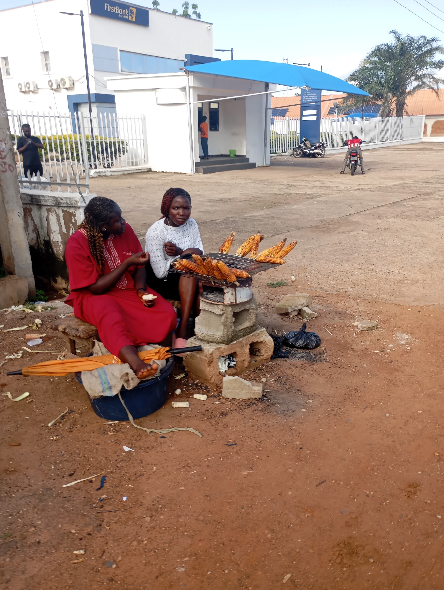 In Shendam, Nigeria, two women are sitting near a roadside stall selling roasted corn. One woman, wearing a red dress, is sitting on a wooden stool and holding a piece of corn. The other woman, wearing a white top, is sitting next to her and looking at the corn on the grill. A portable grill with corn is in front of them, and a large blue basin is to the left of the red-dressed woman, covered with a beige cloth and an orange umbrella. The environment is dusty with scattered debris and a black trash bag. In the background, a FirstBank building with a blue awning is visible, along with a white fence and some palm trees. Two motorcycles are parked near the building.