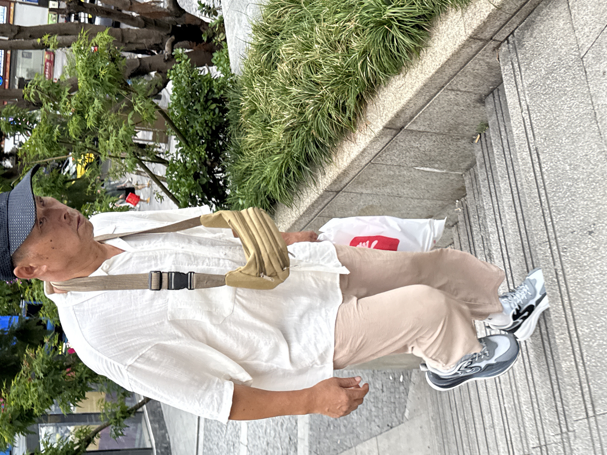 An adult male is walking down a set of outdoor stairs in Yuzhong District, China. He is wearing a white linen shirt, tan pants, a dark baseball cap, and grey and black athletic shoes. A beige cross-body bag is slung over his left shoulder, and he is carrying a white plastic bag in his right hand. Green bushes and trees line the wall to his right. The stairs are made of grey concrete.