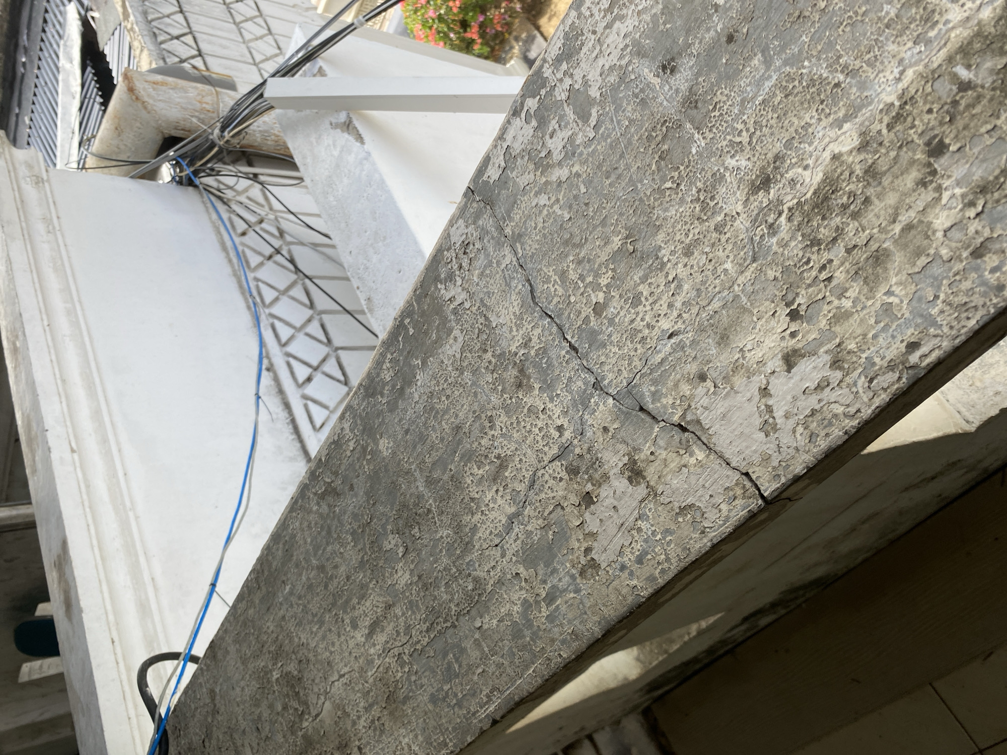 A close-up, outdoor, eye-level shot shows a damaged concrete railing. The concrete surface is textured and weathered, with peeling paint or coating in various shades of gray and off-white. A prominent crack runs diagonally across the railing. In the background, out of focus, are white architectural elements and electrical wires. The scene appears to be in an urban environment, possibly on a balcony or rooftop.