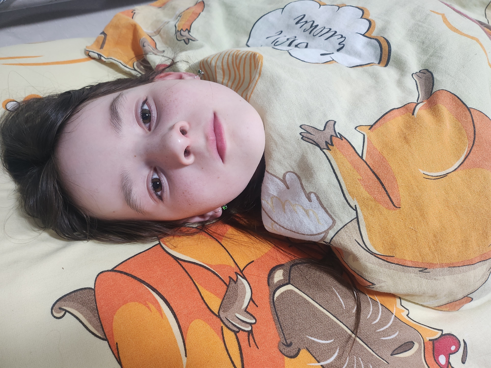 A young girl lies on a bed covered in a cartoon-printed bedsheet. The girl has fair skin, brown hair, and freckles. Her face is tilted slightly upwards, with her eyes looking directly at the camera. She appears to be in a relaxed position. The bedsheet features a pattern of cartoon characters, mainly in shades of orange, brown, and yellow. Based on the surrounding context, it's likely the photo was taken indoors, potentially in a home in Krasnogorskoye, Russia.