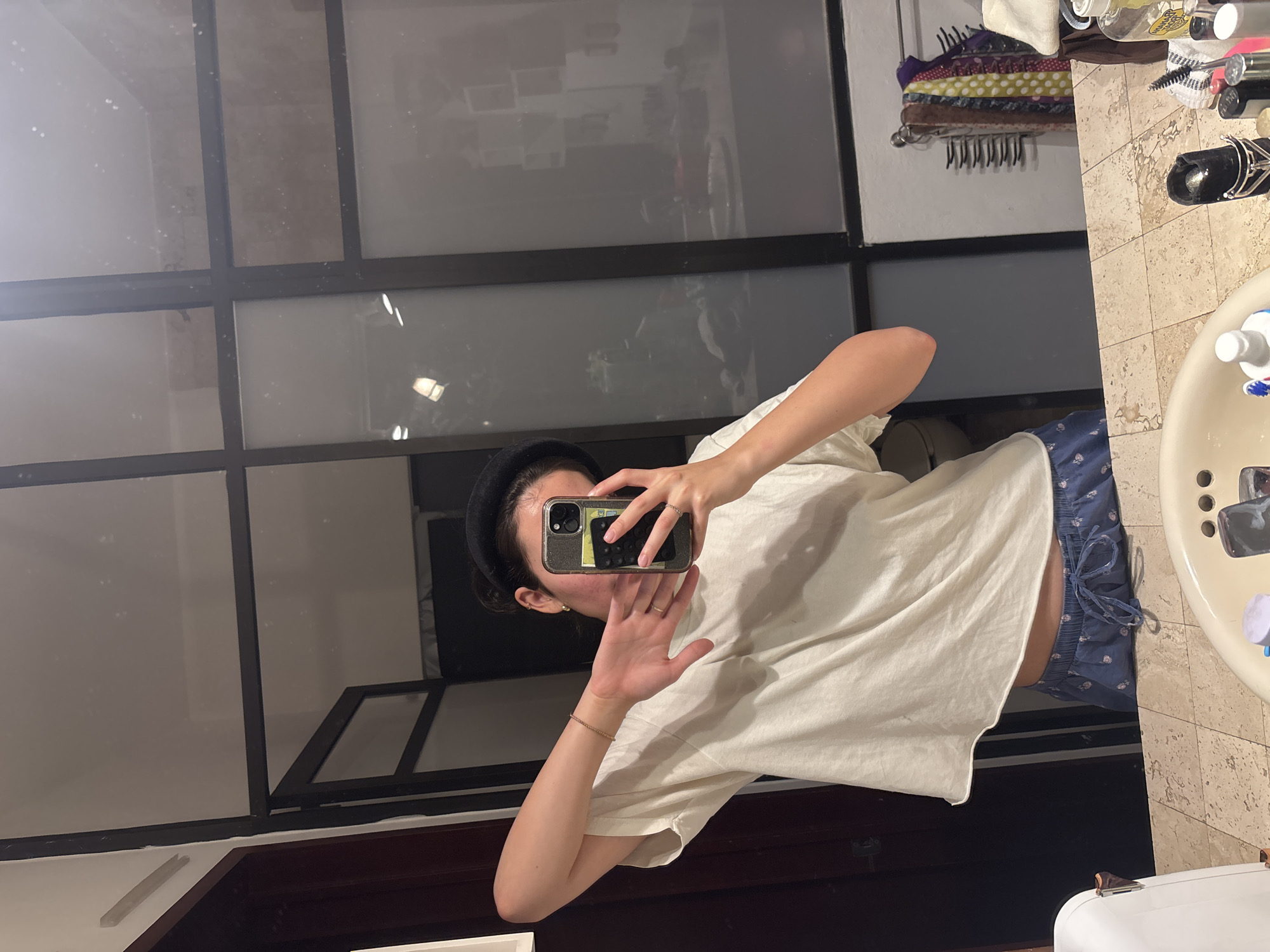 A person wearing a white t-shirt and blue patterned pajama bottoms is taking a selfie in a bathroom. They are holding a smartphone in front of their face, obscuring their features. The bathroom has a tiled floor and sink. A window with multiple panes can be seen in the background, reflecting a cityscape. A rack with hair accessories and various bottles and containers are visible on the counter. It is not possible to pinpoint the specific location within Puebla, Mexico, without more information.