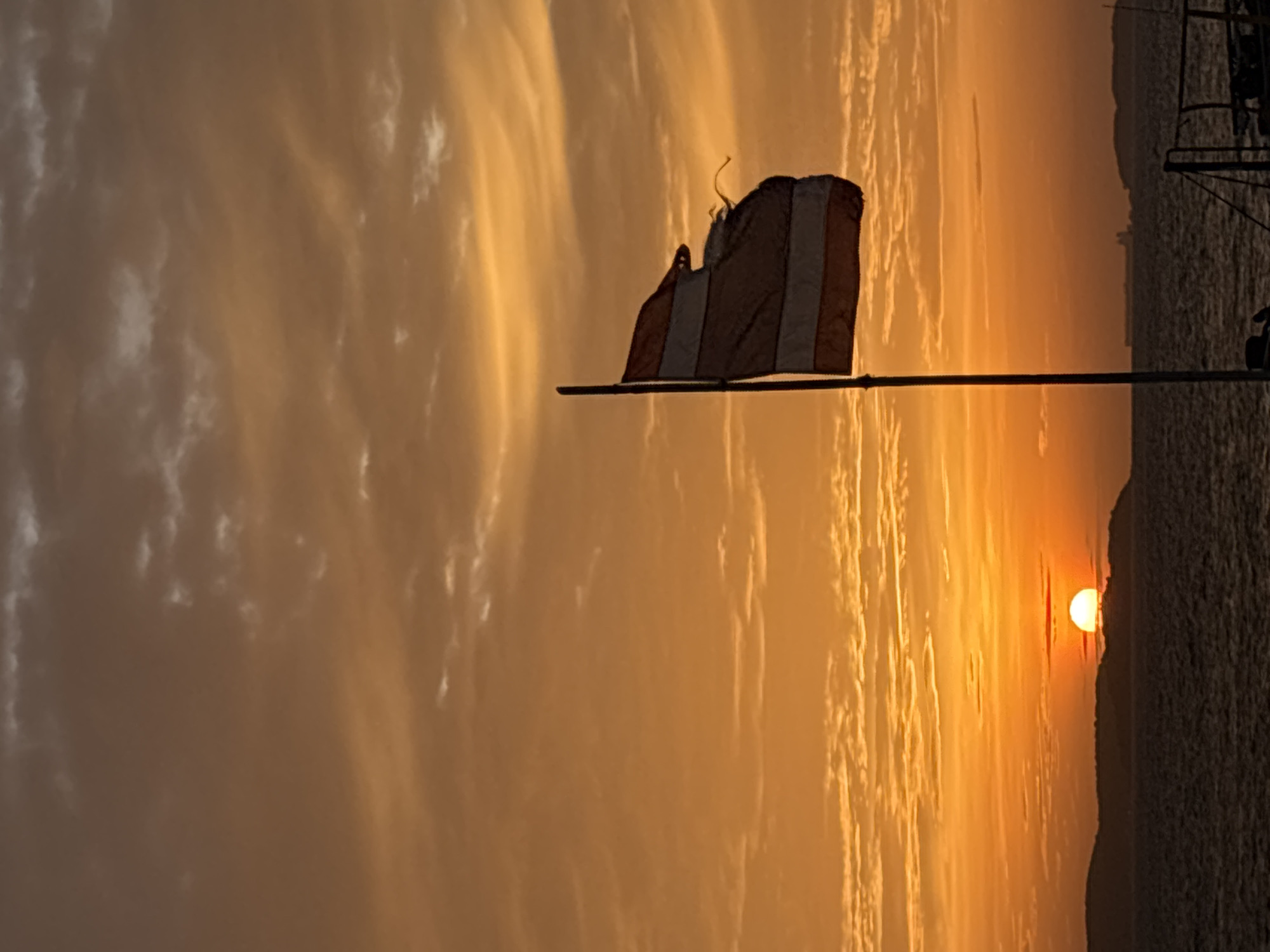 The image captures a serene sunset scene over the sea, likely in Pattaya City, Thailand. A flag, partially silhouetted against the vibrant sky, is prominently featured in the foreground. The flag appears to have alternating stripes of red and white, with a curved element at the top, possibly suggesting a pennant or a unique flag design. It is attached to a horizontal pole, which extends across the frame, creating a stark contrast with the soft, atmospheric clouds.
The sky is filled with dramatic, wispy clouds painted in hues of orange, yellow, and gold, indicative of a sunset. The sun itself is a glowing orb, partially visible and casting a warm light across the horizon. The sea is visible in the background, a dark expanse reflecting the sky's colors. To the right, a dark silhouette suggests land or a structure, possibly a hillside or part of a larger vessel, adding depth to the scene. The overall mood is peaceful and tranquil, evoking a sense of calm as day transitions into night. The time of day is clearly sunset, and the weather appears clear with beautiful cloud formations. There is no visible text in the image.