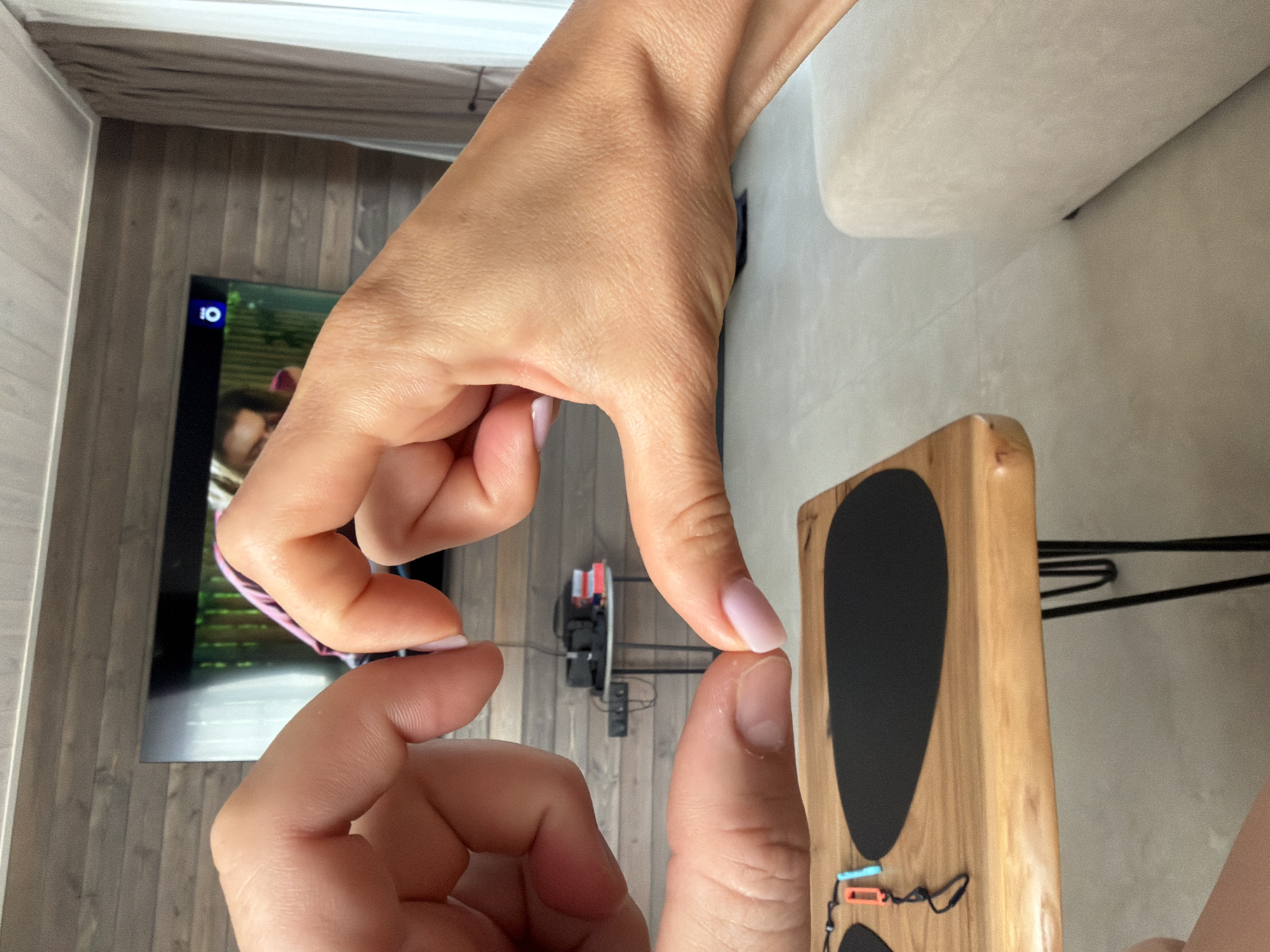 Two hands are forming a heart shape in the foreground. The hands are close together, with the thumbs and index fingers touching to create the top curves of the heart, and the other fingers extending downwards to form the point. The skin tone is light, and the fingernails on the right hand are painted with a light pink polish. In the background, a television screen is visible, showing a person's face and part of a pink garment. To the left of the television, a wooden wall paneling is present. A white curtain hangs on the right side of the image. Below the hands, a wooden table with black hairpin legs is visible. On the table, there are two black oval placemats and a pair of Nintendo Switch controllers, one orange and one blue, attached by a black strap. The floor is a light, neutral color. A portion of a light-colored sofa is visible on the far right of the frame. The image appears to have been taken indoors, possibly in a living room. The location is not explicitly stated in the image, therefore no information about Zarozhdeniye, Russia can be provided.