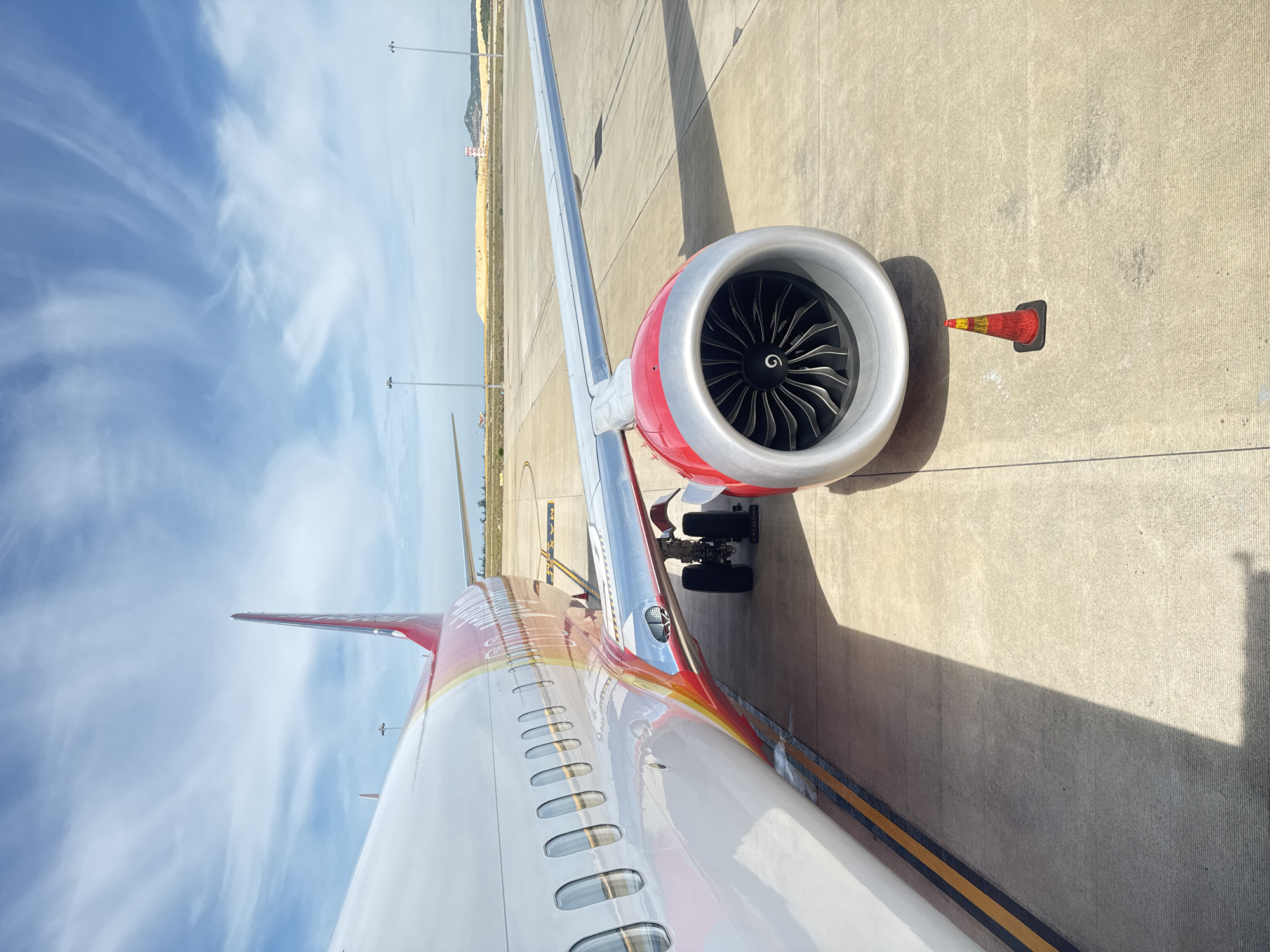 An airplane engine, painted red and silver, is in the foreground on the tarmac at Cam Ranh International Airport in Cam Ranh, Vietnam. The airplane's fuselage, white with red and yellow accents, extends to the left. The wing, also silver, stretches out to the right. A small orange and yellow traffic cone sits on the concrete ground in front of the engine. In the background, a large pile of sand and construction equipment are visible, along with distant hills under a bright blue sky with wispy clouds. The scene is brightly lit by the sun, casting long shadows.
