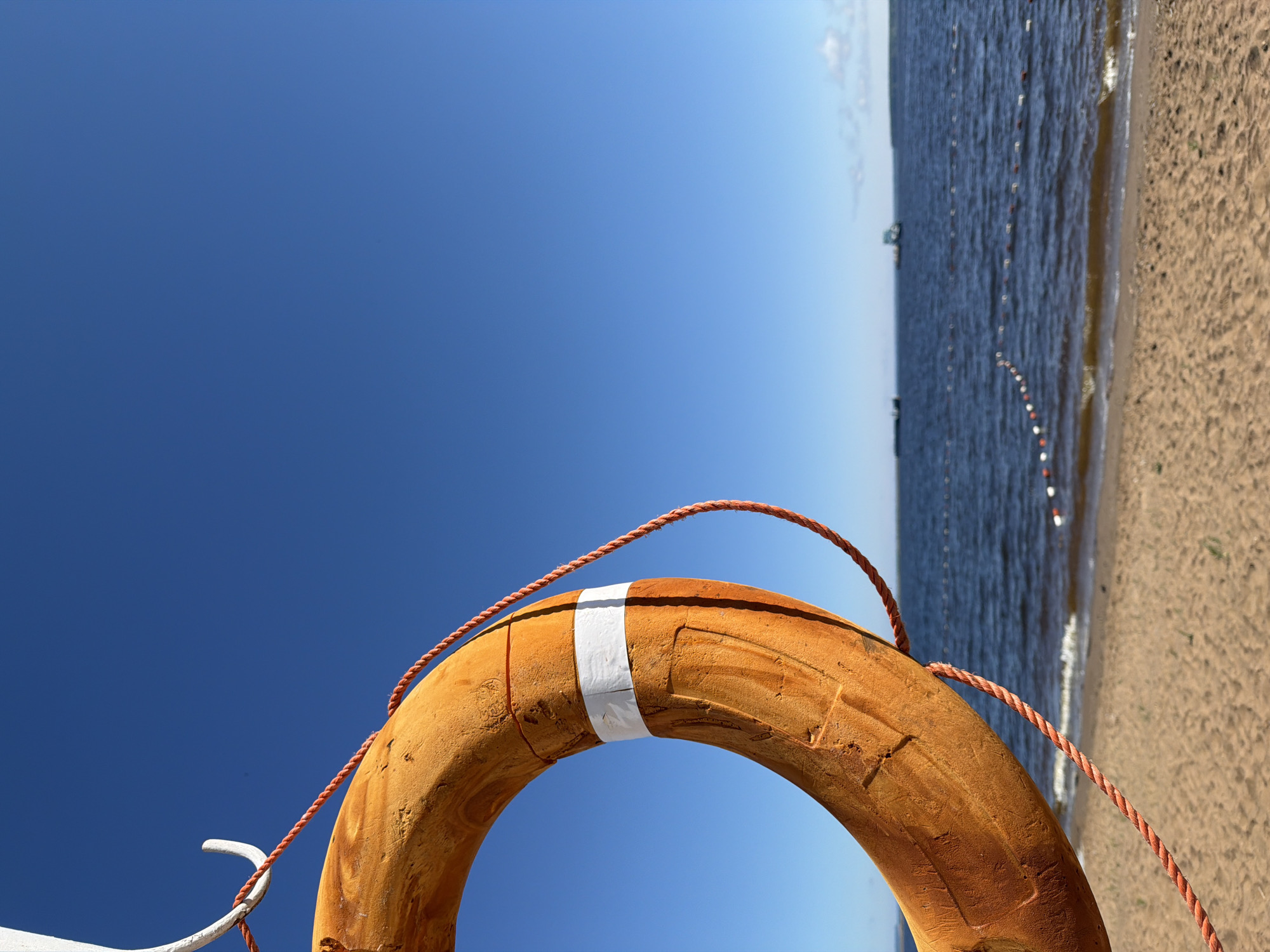 A close-up, low-angle shot features an orange life preserver with a white band, suspended by a white hook and a red rope. The life preserver occupies the left side of the frame, against a clear, vibrant blue sky. In the background, a calm, dark blue body of water stretches to the horizon, where a sandy beach is visible in the foreground. A series of buoys, some red and white, mark a boundary in the water. Two distant boats are visible on the water. This scene is indicative of a waterfront location in Chaikovsky, Russia, likely a beach or swimming area. There are no people present in the image.